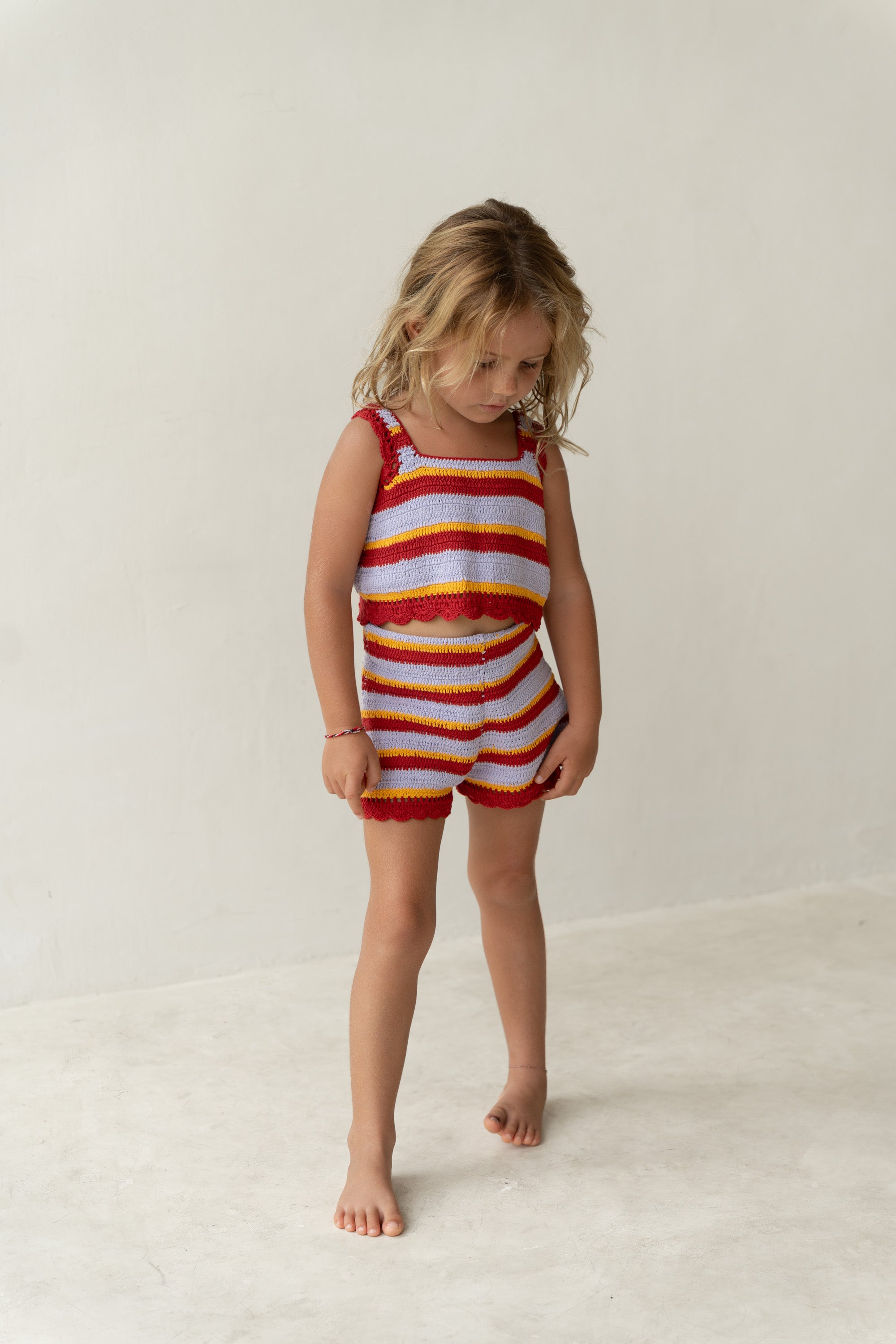 A young child with long blonde hair stands barefoot on a light-colored floor, wearing ILLOURA THE LABEL's Crochet Top Sunset Stripe and matching shorts in vibrant hues of red, blue, and yellow. The background is a plain, light wall.