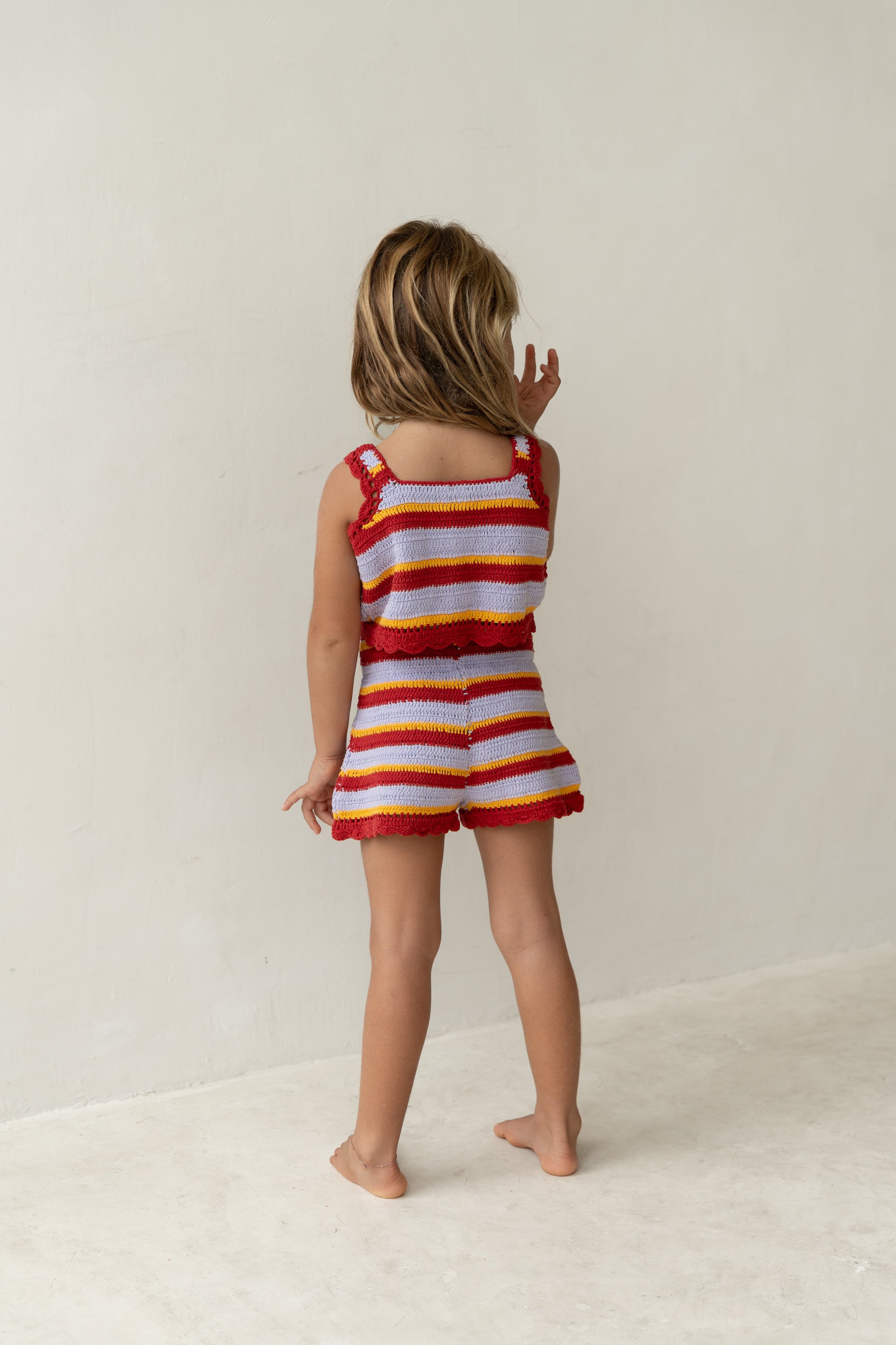 A young child with light brown hair stands barefoot, gazing at a blank wall. They're dressed in a vacation-style, colorful outfit from ILLOURA THE LABEL, featuring Crochet Shorts Sunset Stripe with Retro Sunset hues in red, yellow, and white horizontal patterns.