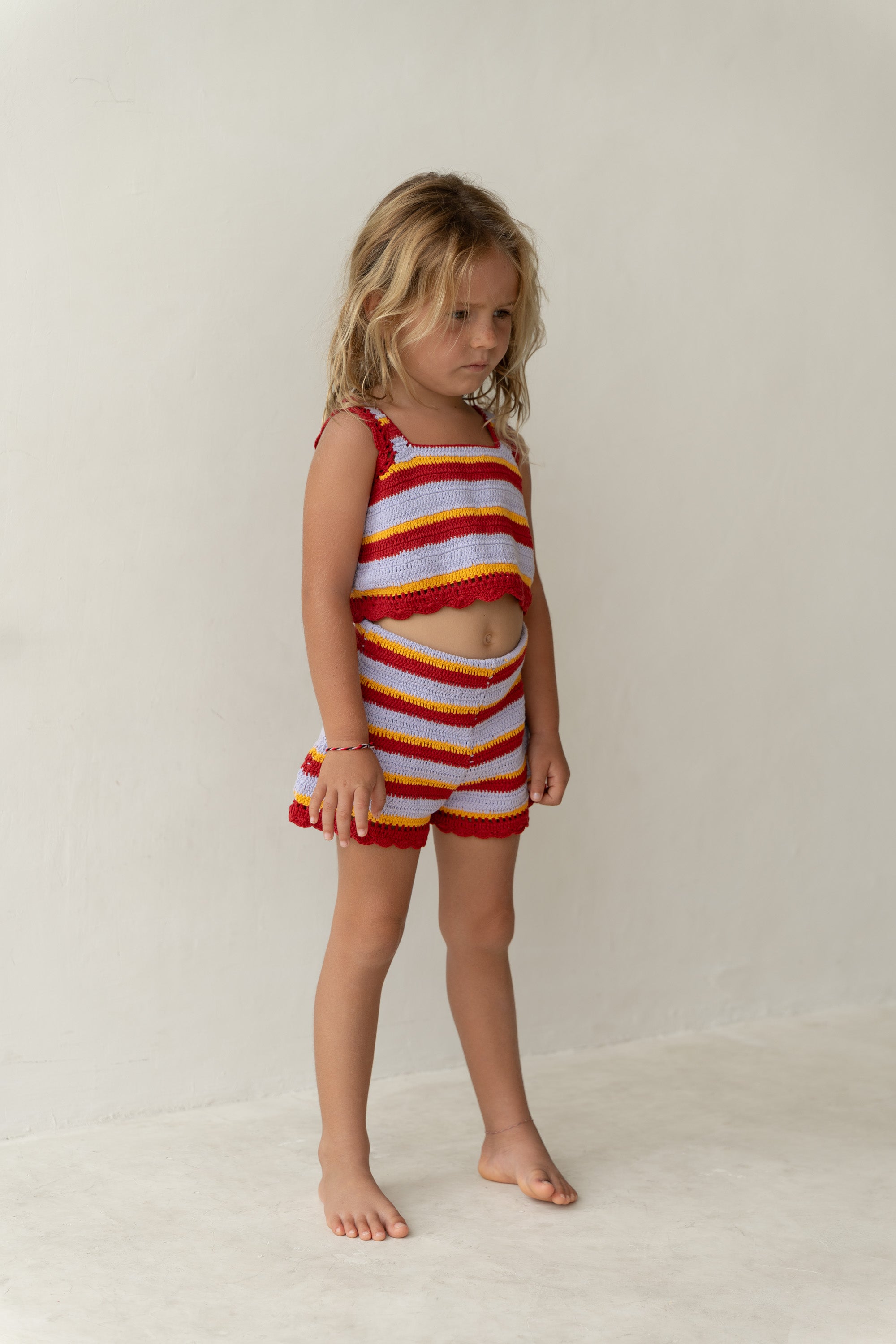 A young child stands barefoot against a plain backdrop, adorned in ILLOURA THE LABEL's Crochet Top Sunset Stripe, an outfit featuring sunset hues of red, orange, and white. The ensemble is made with 100% cotton crochet and complements the child's long, wavy hair and neutral or slightly pensive expression.