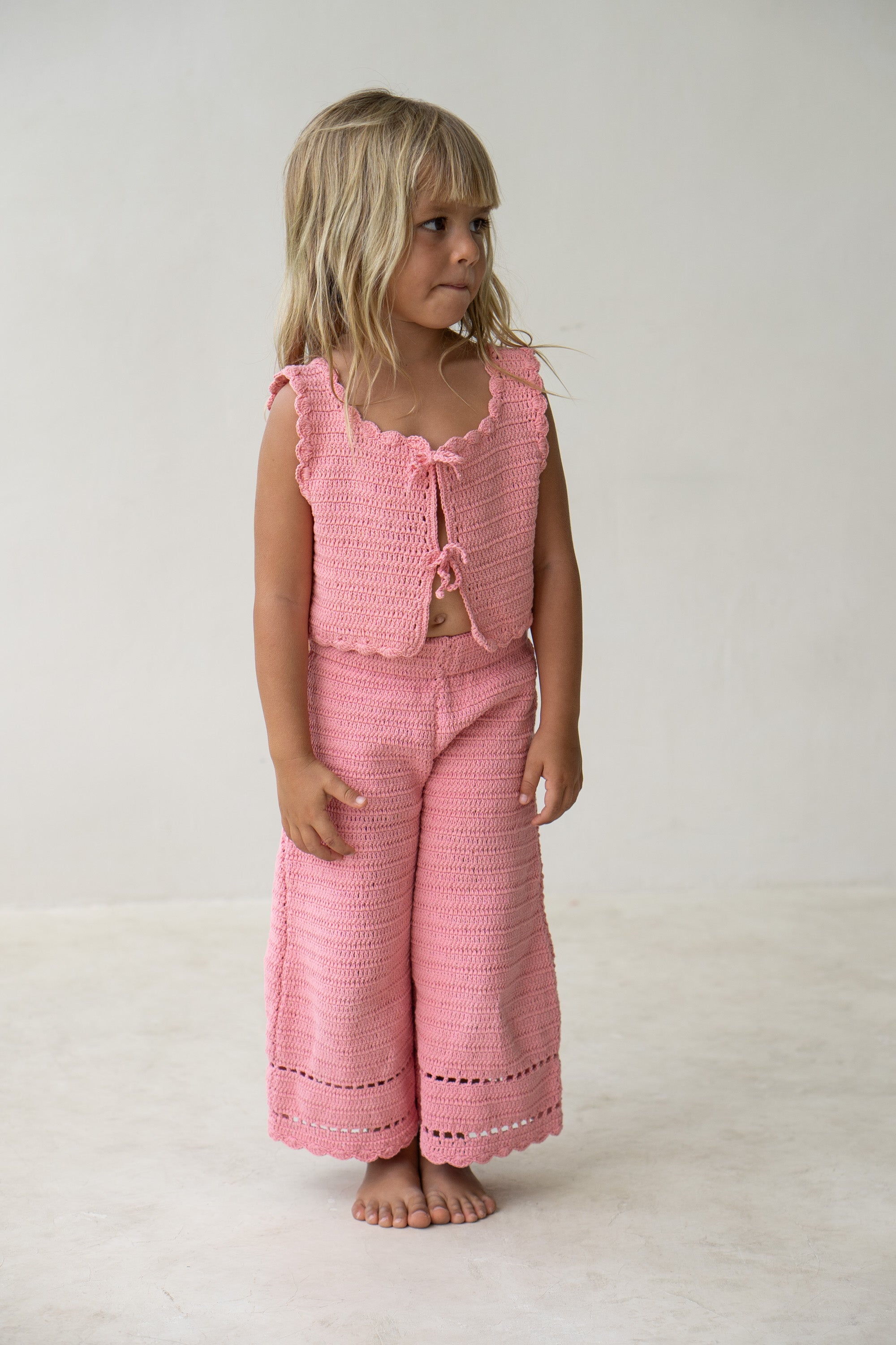 A young child with long blond hair stands barefoot against a plain light gray background. They are dressed in ILLOURA THE LABEL's "Crochet Pants Coral" outfit, which includes a sleeveless pink knit top adorned with ruffles and a bow at the chest, paired with matching wide-legged pants featuring an elasticated waistband. They look slightly to the side.