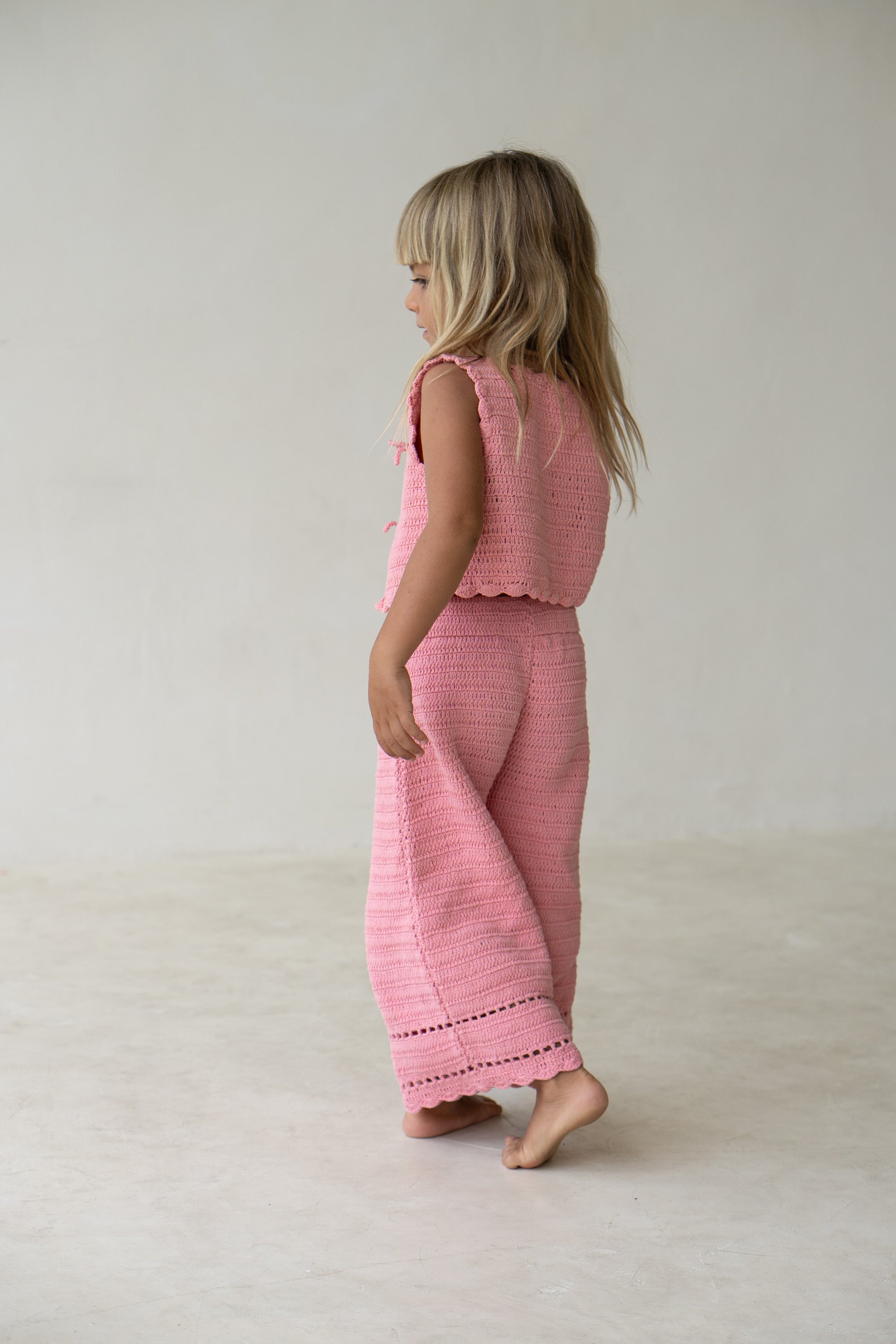 A young child with long, blonde hair is facing away and walking barefoot. They are wearing ILLOURA THE LABEL's Crochet Vest Coral in a delicate cotton crochet design, paired with matching pants. The background features a plain, light-colored wall and floor.