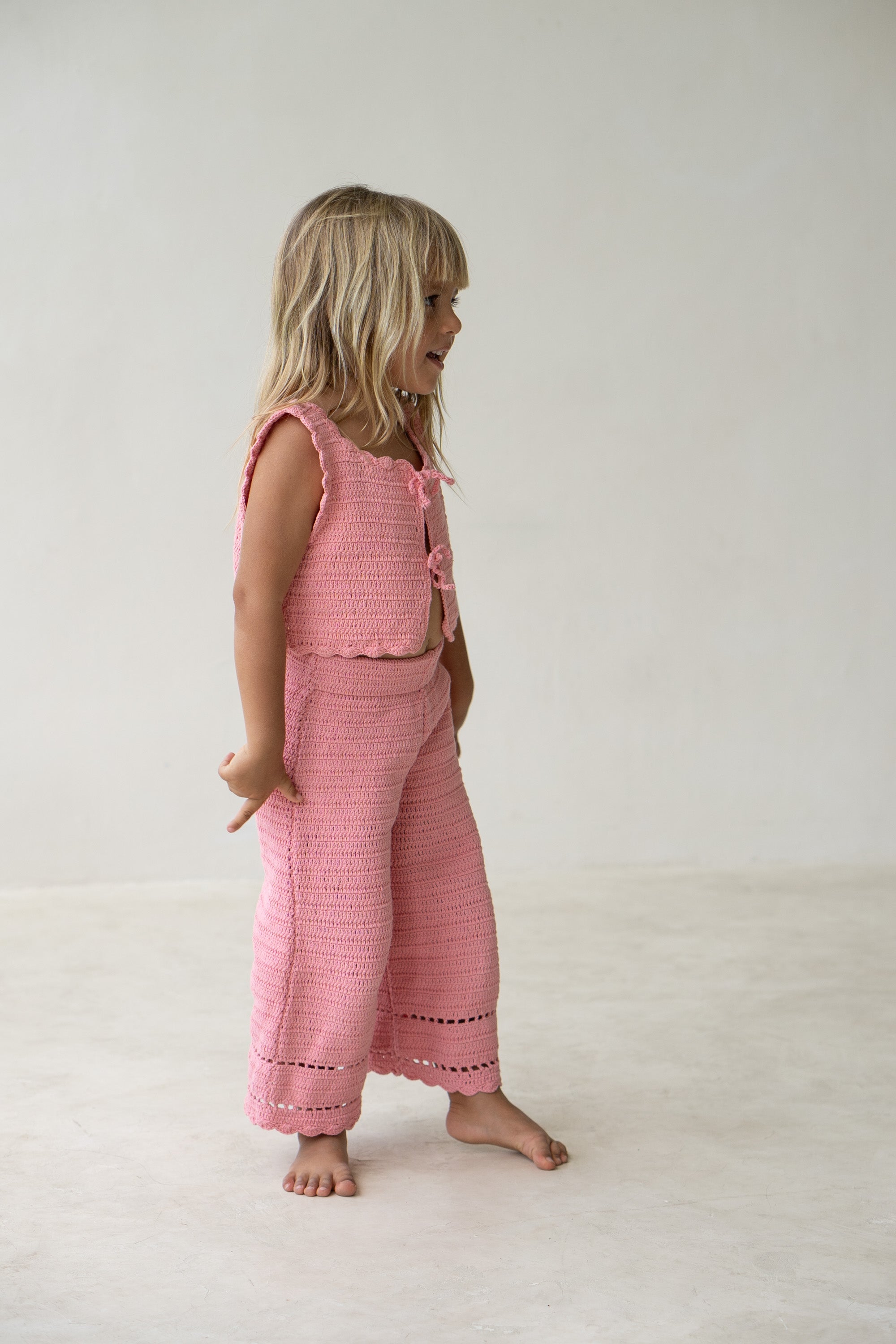 A young child with long, blonde hair stands barefoot on a light-colored floor. They are wearing a matching set in a coral colourway, featuring the Crochet Vest Coral from ILLOURA THE LABEL and wide-leg pants. Looking to the side with their hands behind their back, they exude effortless charm.