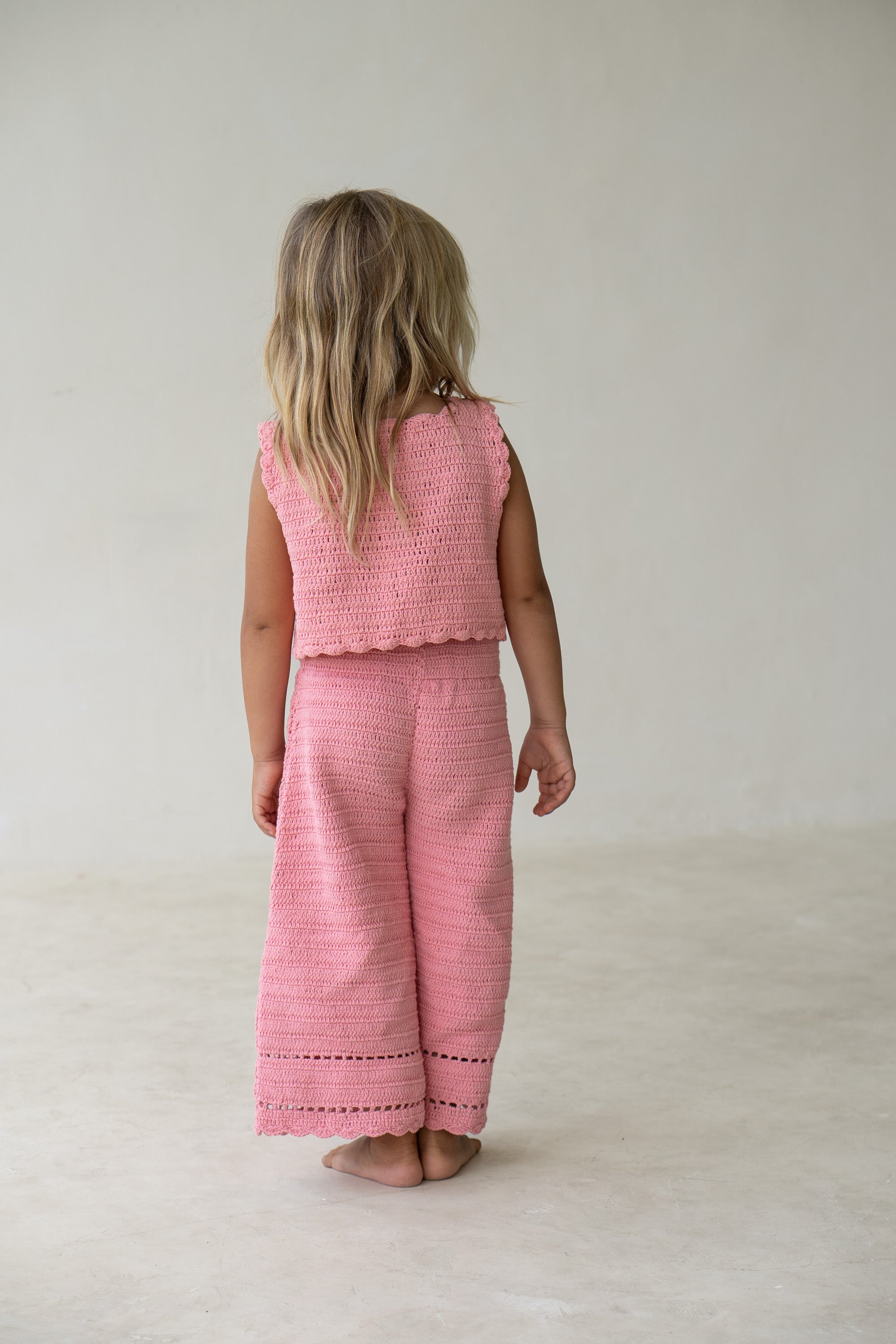 A young child with long blond hair, seen from behind, stands barefoot on a light-colored floor. They are wearing the Crochet Vest Coral and matching wide-legged pants from ILLOURA THE LABEL. The background is plain and light-colored.