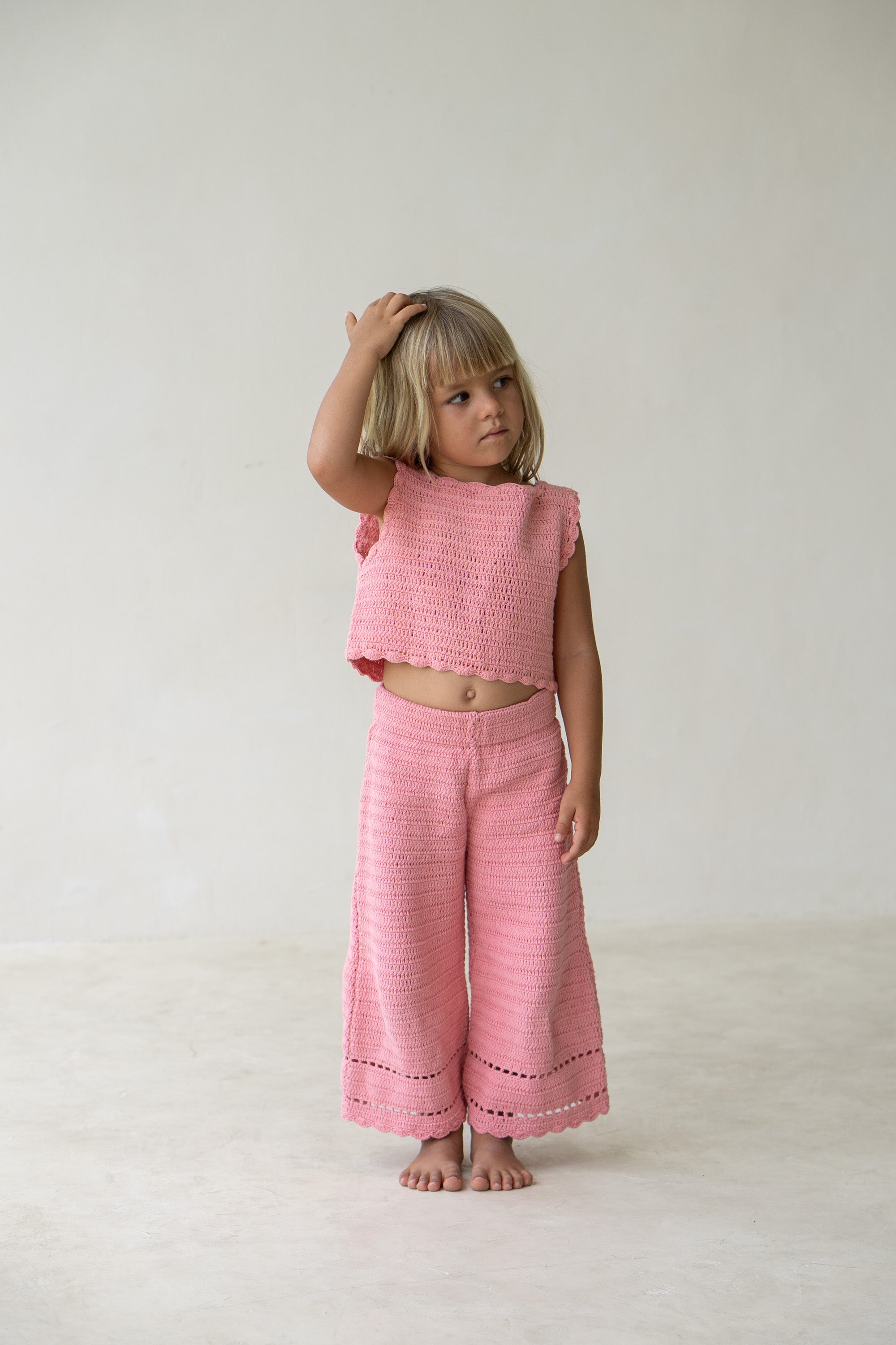 A young child stands barefoot on a light-colored floor, wearing the Crochet Pants Coral and a matching pink cotton crochet top from ILLOURA THE LABEL. The child has short blonde hair and is looking to the side, with one hand touching their head. The background is a plain off-white wall.