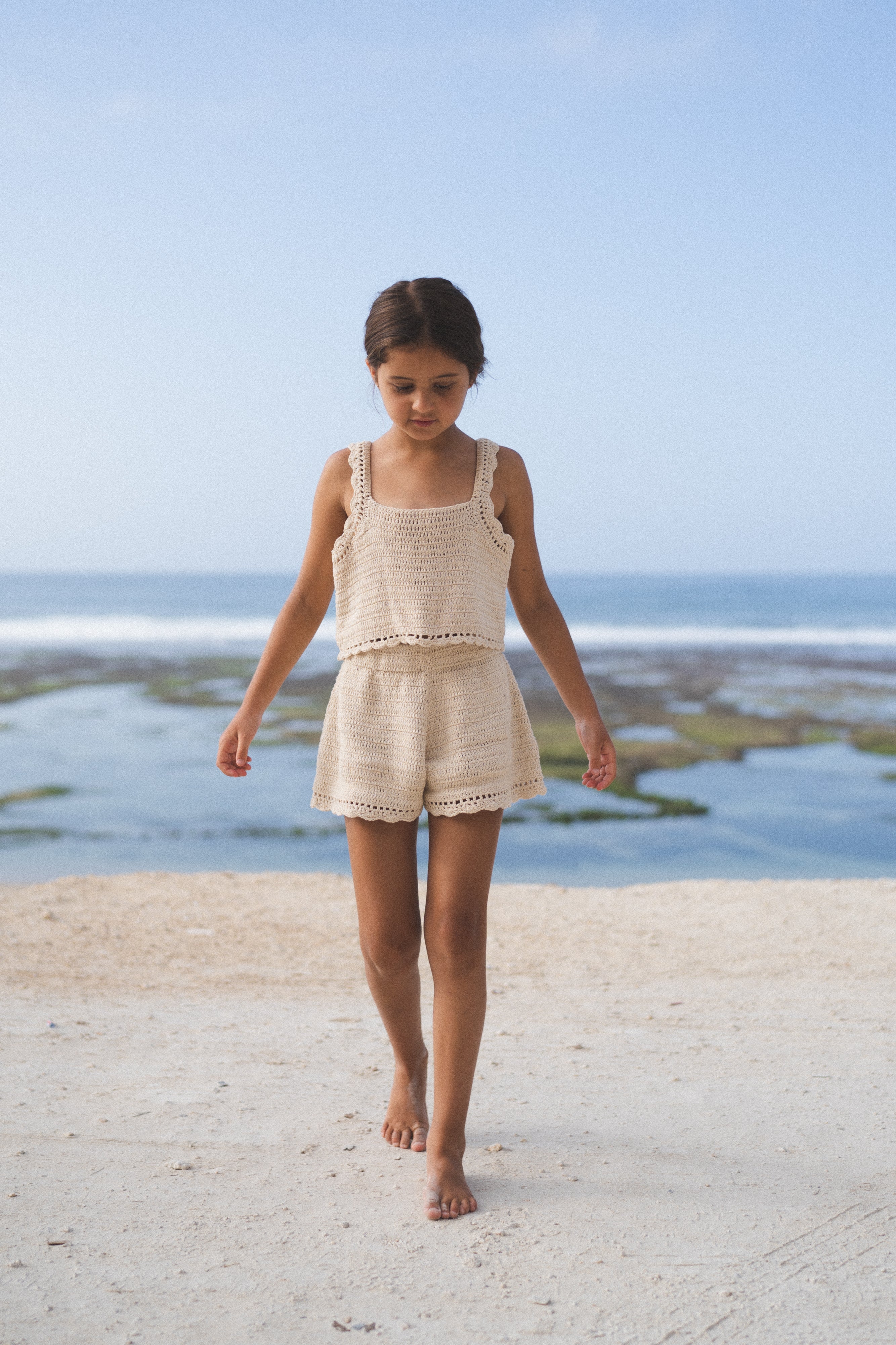 A young girl wearing the ILLOURA THE LABEL Crochet Top Ecru and matching shorts walks barefoot on sandy ground, with the ocean and a clear blue sky in the background.