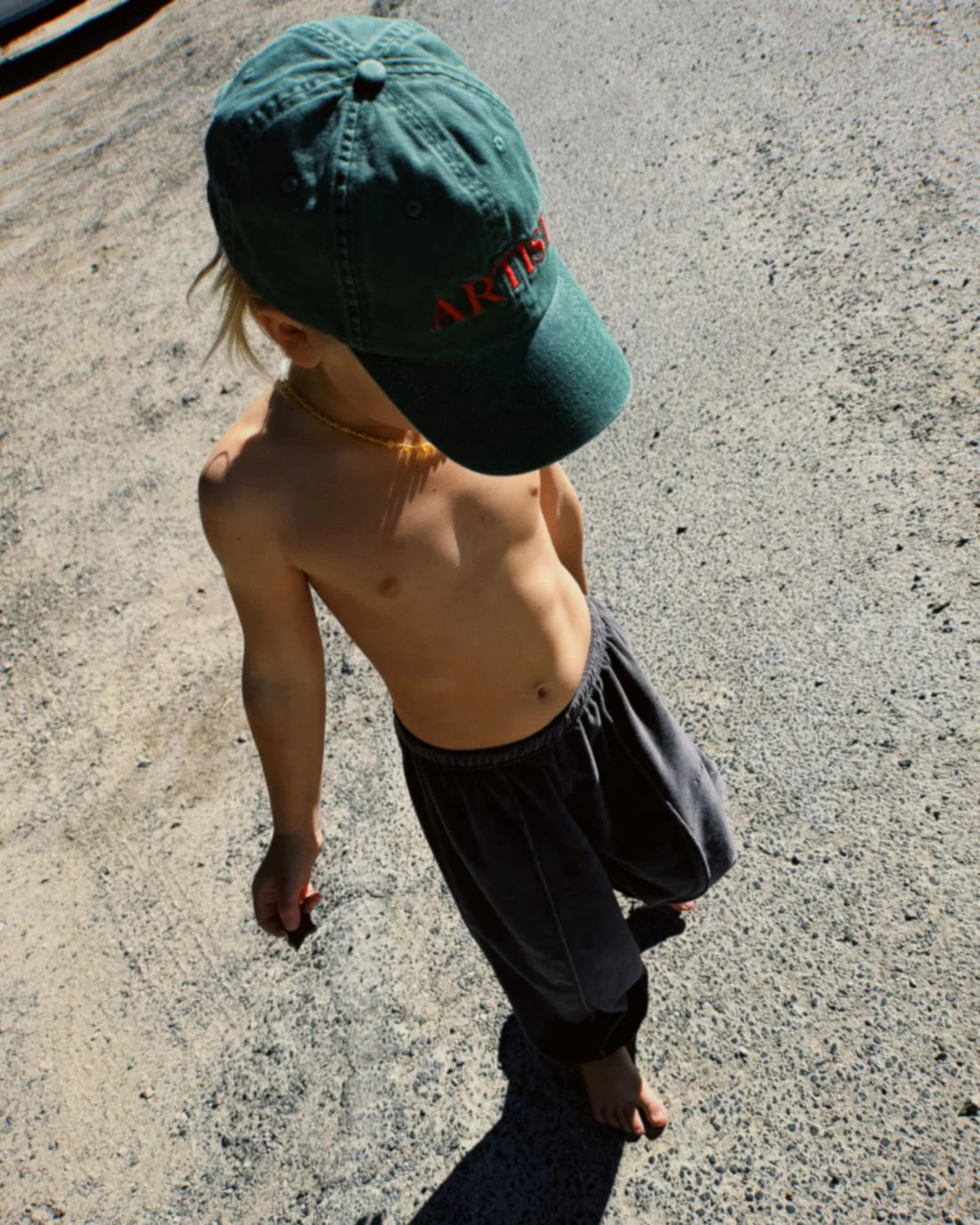 A child with long hair, wearing a Ziggy Zaza ~ Artist Cap Forest Green and dark pants, is walking barefoot on a sunlit concrete surface.