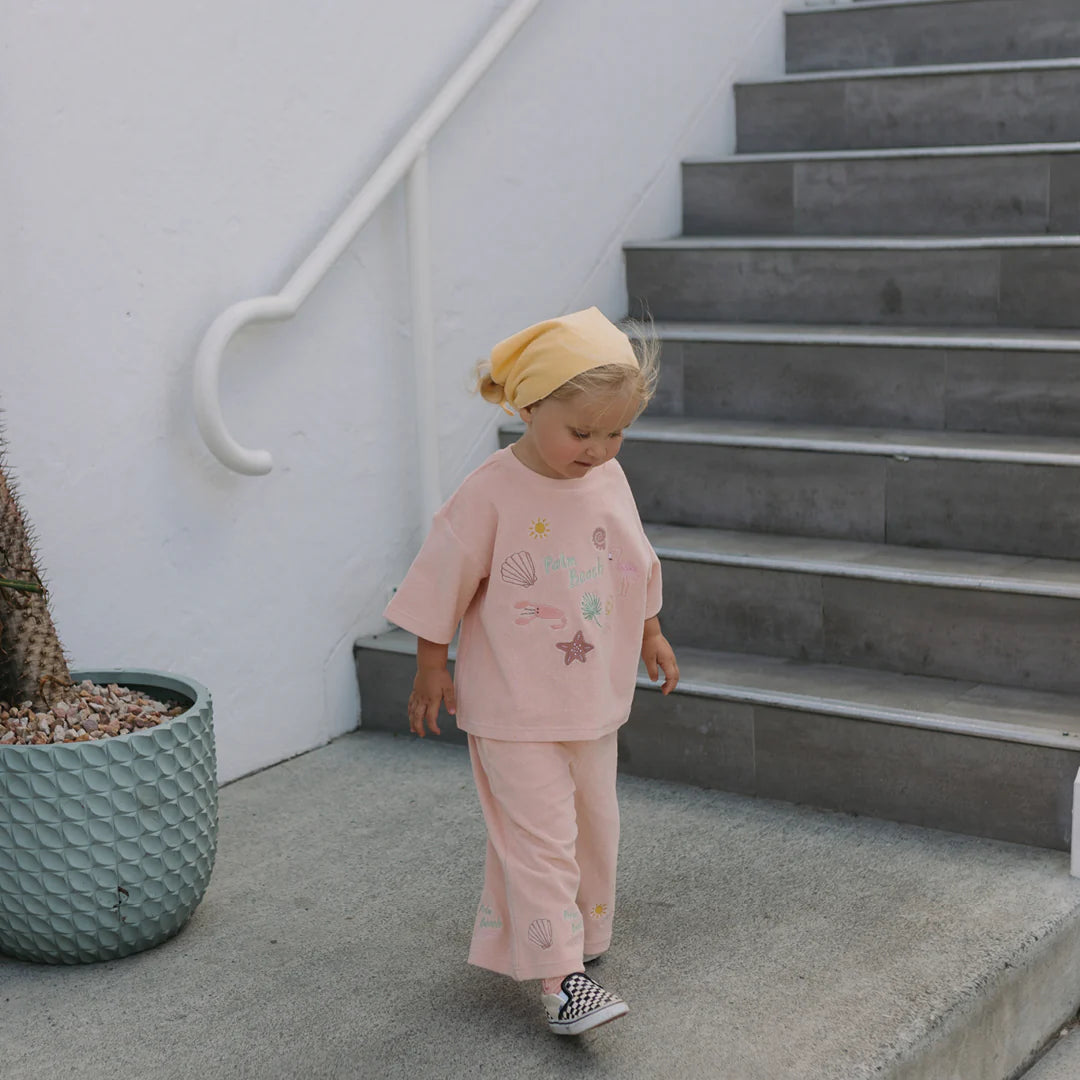 A young child wearing the Palm Beach Wide Leg Pants Terry Towel in Flamingo Pink by GOLDEN CHILDREN walks down gray stairs. The light pink outfit features sea-themed designs and is complemented by a yellow headscarf. The soft terry towel cotton fabric drapes elegantly as the child passes by a cactus in a large textured planter, set outdoors beside a white wall with a railing.