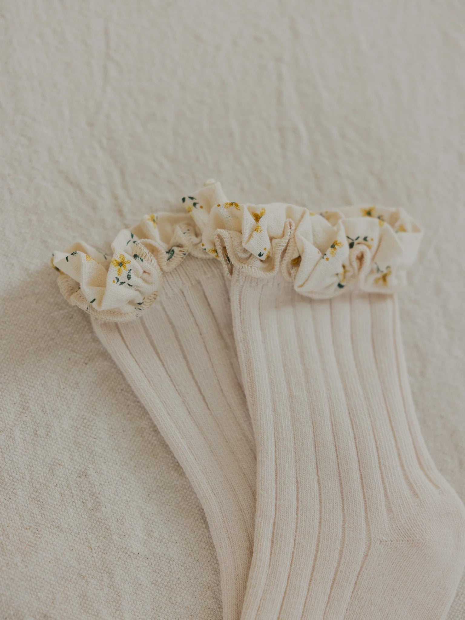 A close-up shot of the Marigold Socks Sand by VALENCIA BYRON BAY, showcasing their creamy-white hue and frilly top adorned with delicate floral patterns in shades of yellow and green. The socks are placed on a textured, light-colored fabric background that evokes the charming textiles found in Byron Bay.