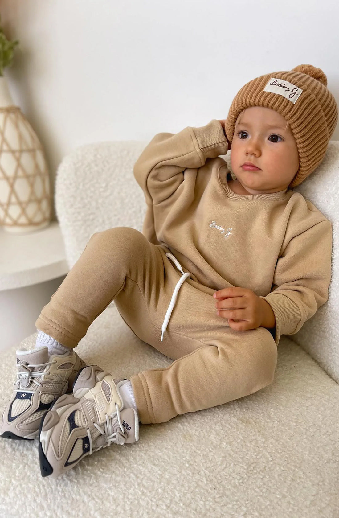 A young toddler wearing the pom pom beanie in rusty with a matching light brown jumper and pant set and sneakers.