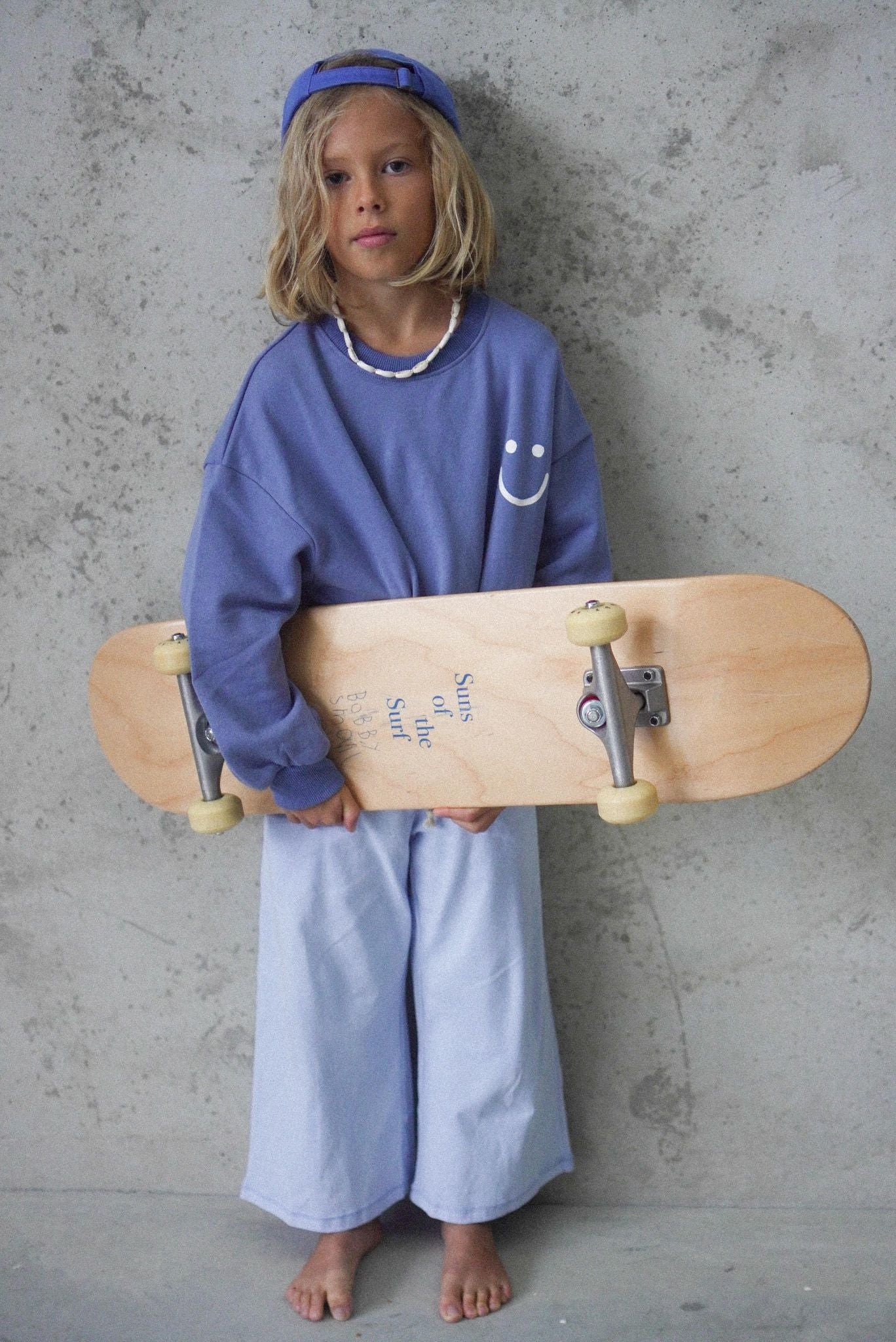 Young boy wearing the wave of life blue pullover and a skateboard and wearing a blue hat and pants.