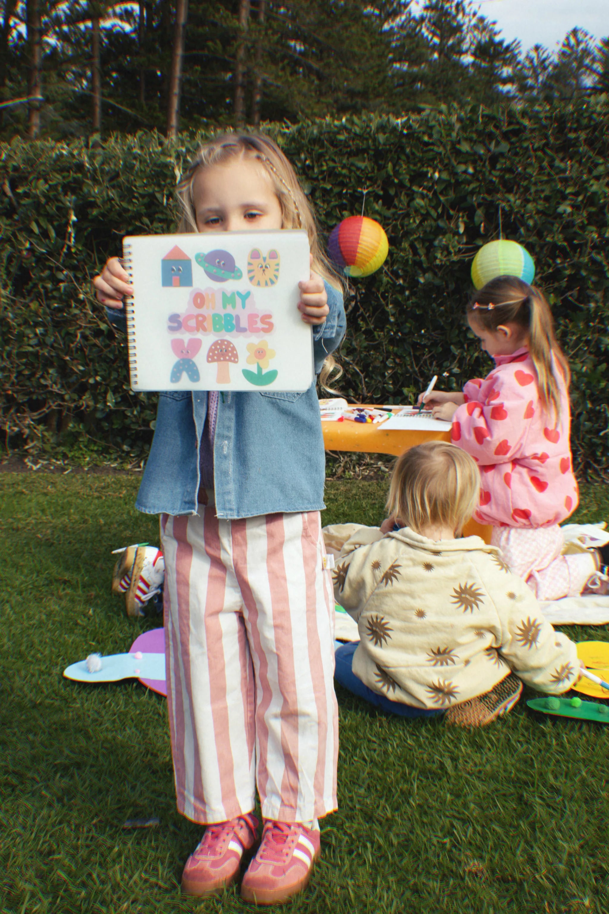 On the grass, a child proudly shows an artwork titled "Oh My Scribbles" from the OH MY FRANKIE reusable coloring book. Behind, two kids craft on a picnic blanket, surrounded by hedges and lanterns that add to the playful and imaginative vibe.