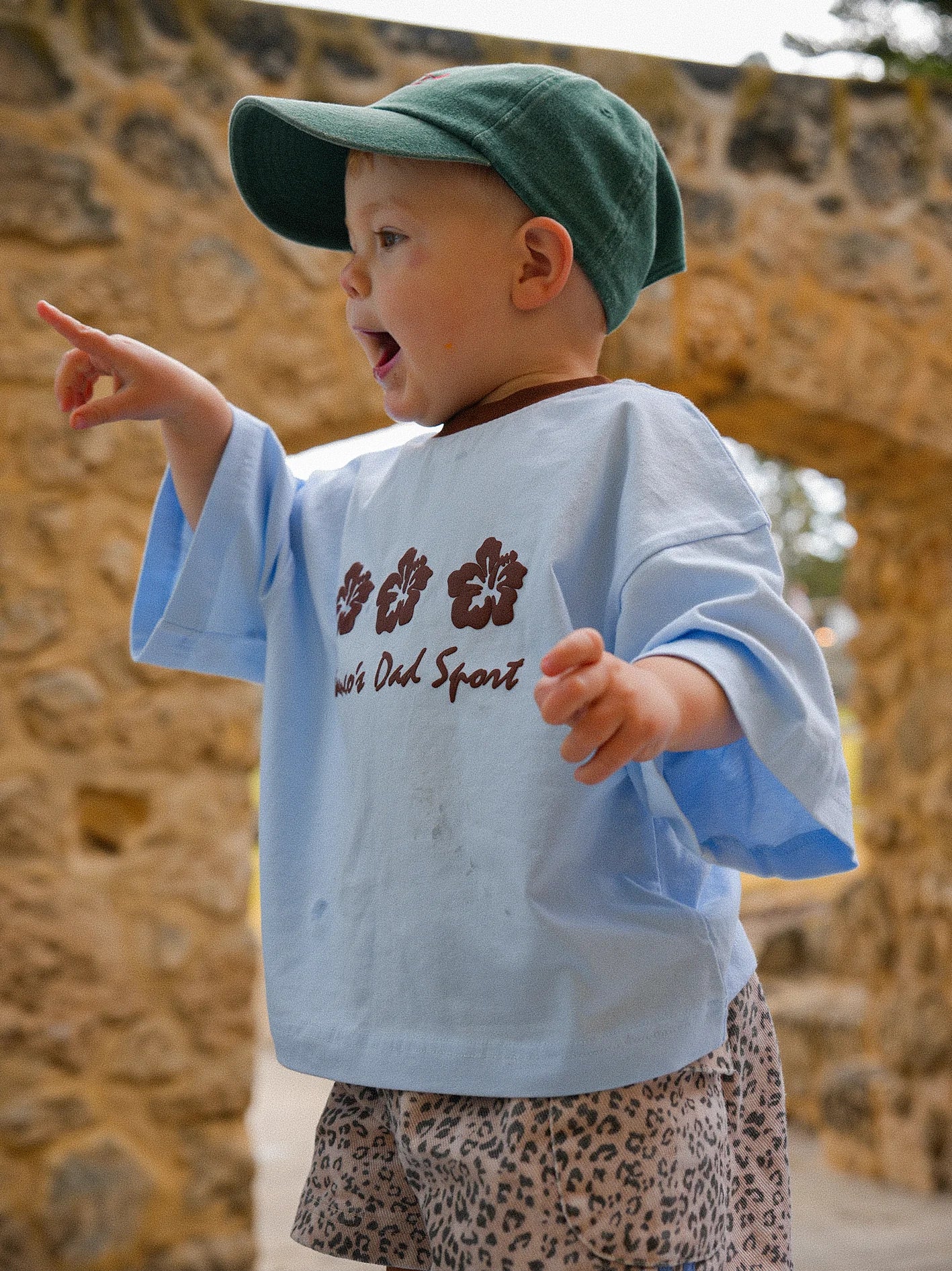 A toddler wearing the PRE-ORDER Hibiscus Tee by FRANCO'S DAD, paired with leopard print shorts and a green cap, points enthusiastically at something off-camera. The child is standing in front of a textured stone wall with an archway.