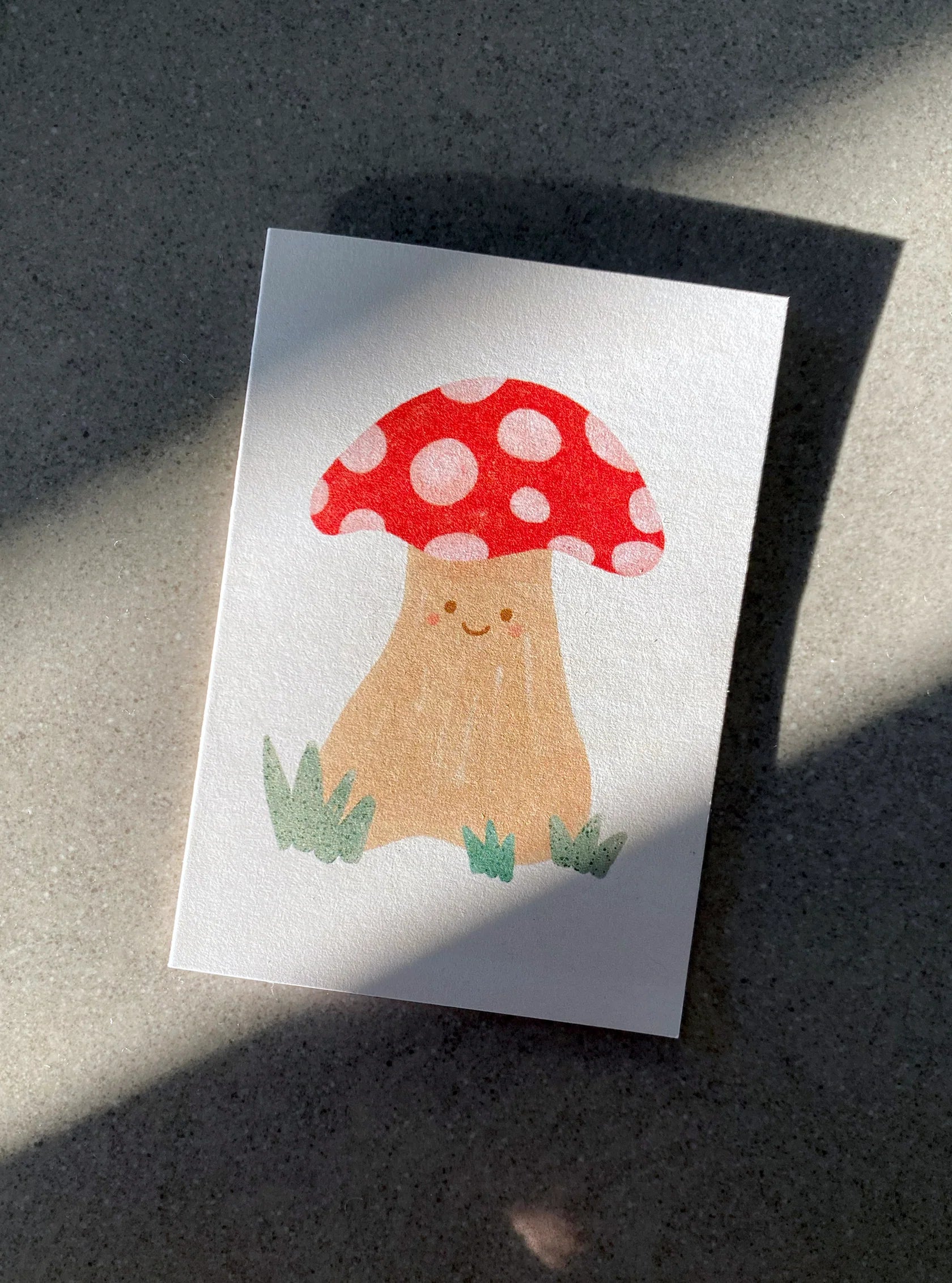 This adorable Mushroom Mini Card by LAUREN SISSONS STUDIO features a charming illustration of a smiling mushroom with a red cap adorned with white spots. The mushroom has a beige stem standing on a tuft of grass. Made from recycled paper, the card is displayed on a flat surface, where shadows cast intricate light streaks across it.