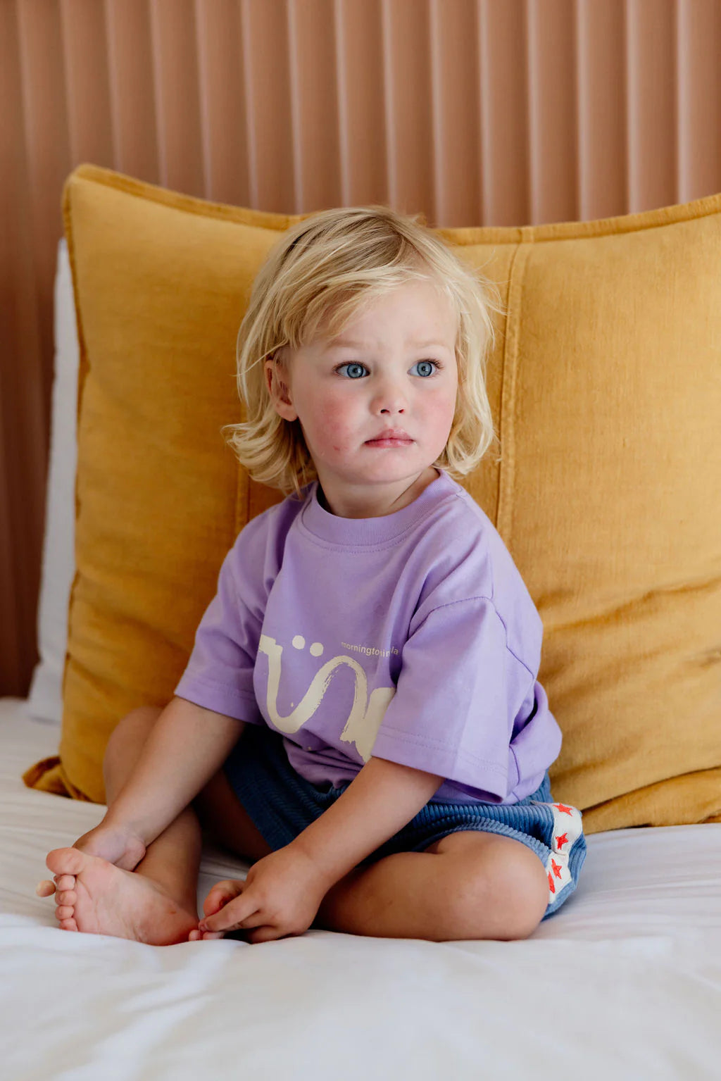 A young child with blond hair sits on a bed against mustard yellow pillows, wearing an oversized Sunday Siblings ~ Il Sole Tee Purple/Cream and denim shorts, looking to the side with a neutral expression.