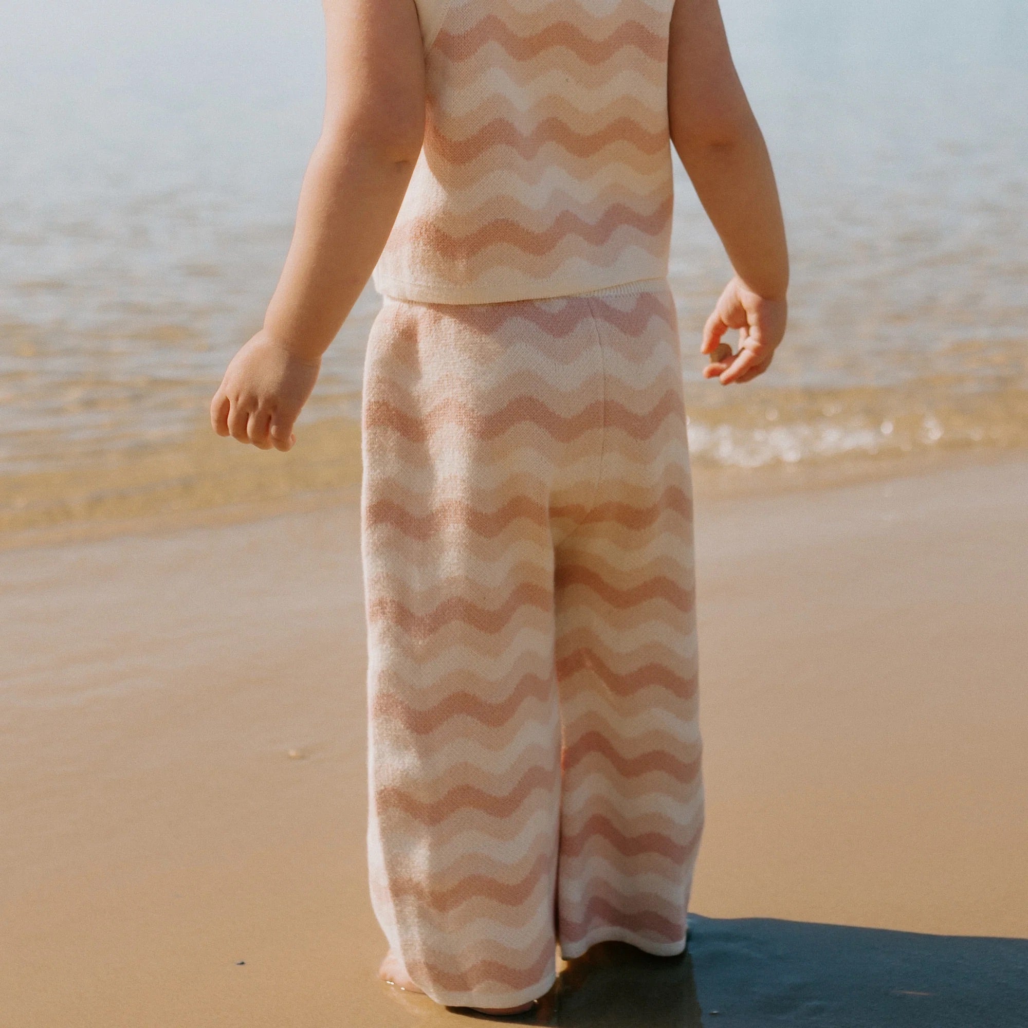 A child wearing the Seashore Knit Set Strawberry Cream from GOLDEN CHILDREN, featuring a relaxed fit and sleeveless top with wide-legged pants in a wavy pink and white pattern, stands on a sandy beach facing the water. The seaside-inspired attire radiates warmly under the sun's light.