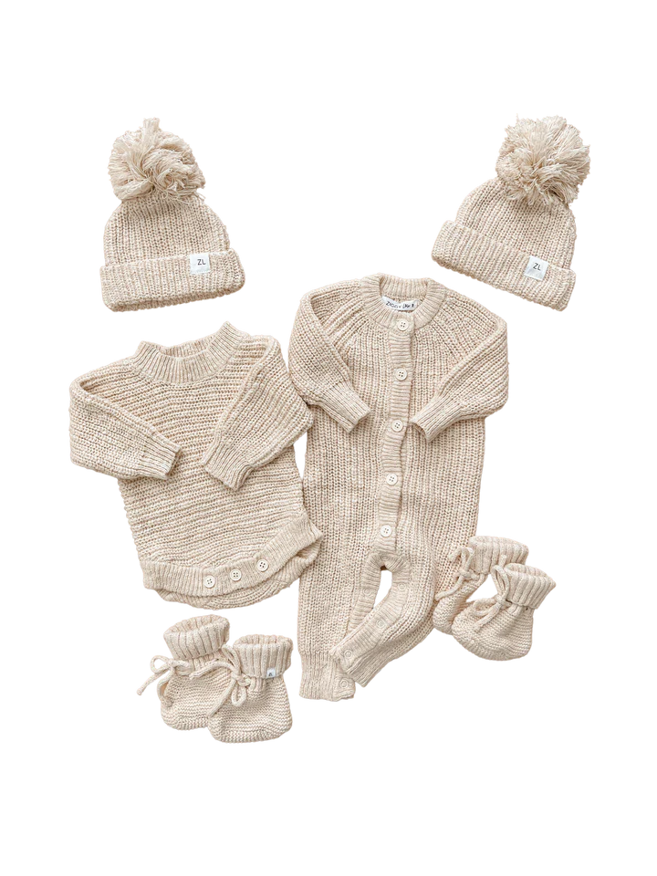 A set of beige knit baby clothing from the Ziggy Lou Heirloom Honey Collection includes two beanies with pom-poms, a button-up knitted romper named "Heirloom Romper Honey," a sweater with a long back, two pairs of booties, and two pairs of mittens. The items are neatly arranged on a white background.