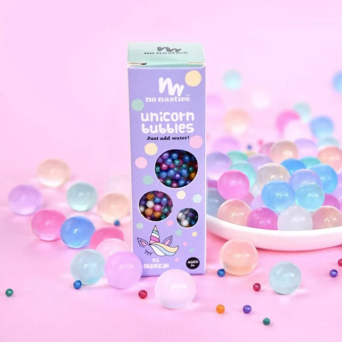 A package labeled "Unicorn Bubbles Pastel Biodegradable Water Beads" by NO NASTIES, showcasing a purple design, is set against a gradient background of pink and purple. It's surrounded by vibrant water beads in hues of pink, blue, and purple. Ideal for sensory play, the box is adorned with a small unicorn illustration that encourages fine motor skills exploration.