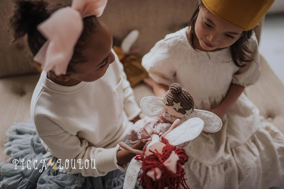 Two young girls sit on a couch, smiling and gazing at each other while holding PICCA LOULOU Fairy Celeste 35cm dolls. One girl wears a large pink bow, and the other sports a mustard yellow headband. The background is softly blurred, drawing attention to the girls and their cherished dolls. Text on the image reads "PICCA LOULOU.