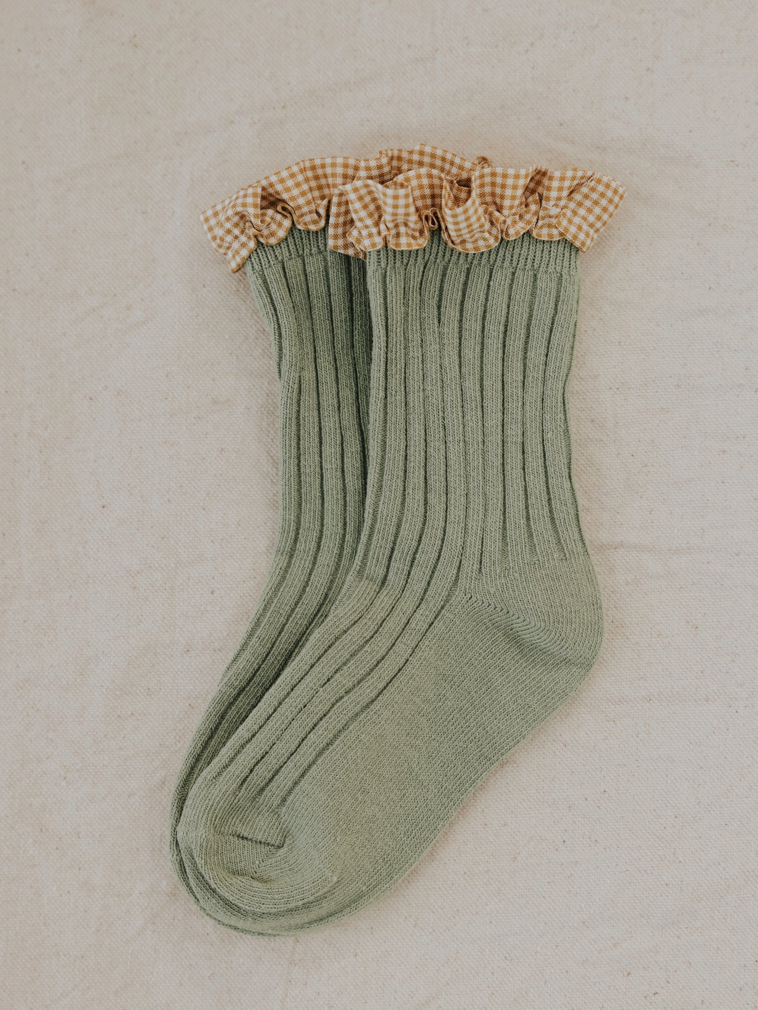 Frilly Socks Avocado from VALENCIA BYRON BAY, in a green ribbed cotton design with a checkered ruffle at the top, lying on a neutral-colored fabric background. The ruffle features a classic white and beige gingham pattern, adding a playful touch to this cozy frilly socks design.