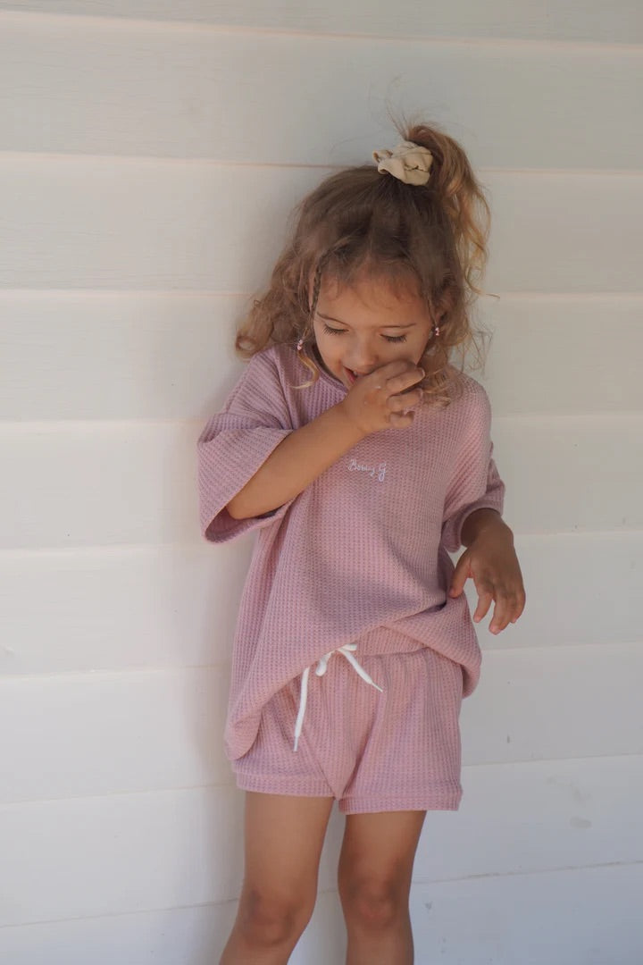 A young child with curly hair, wearing BOBBY G BABY WEAR's Coby Shorties Dusty Rose, smiles while covering their mouth. The soft waffle fabric outfit with a stretchy waistband ensures comfort against a plain light-colored wall, enhancing this charming scene.