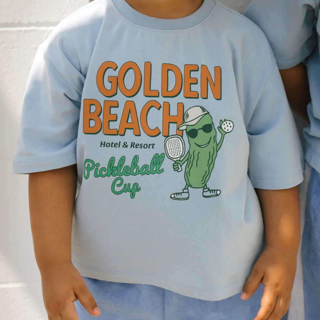 Two children wear comfortable, palm blue mid-sleeve tees from GOLDEN CHILDREN. One of the shirts features a playful pickle character with a paddle and ball, accompanied by the text "Golden Beach Hotel & Resort Pickleball Cup" in vibrant orange and green.