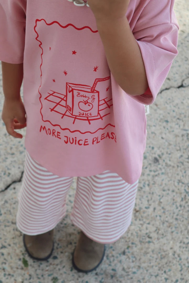 A child wearing BOBBY G BABY WEAR's Floaty Pants - Candy Cane Pink and brown boots sports an organic cotton shirt with a juice box image and "More Juice Please" in red on a textured concrete surface.