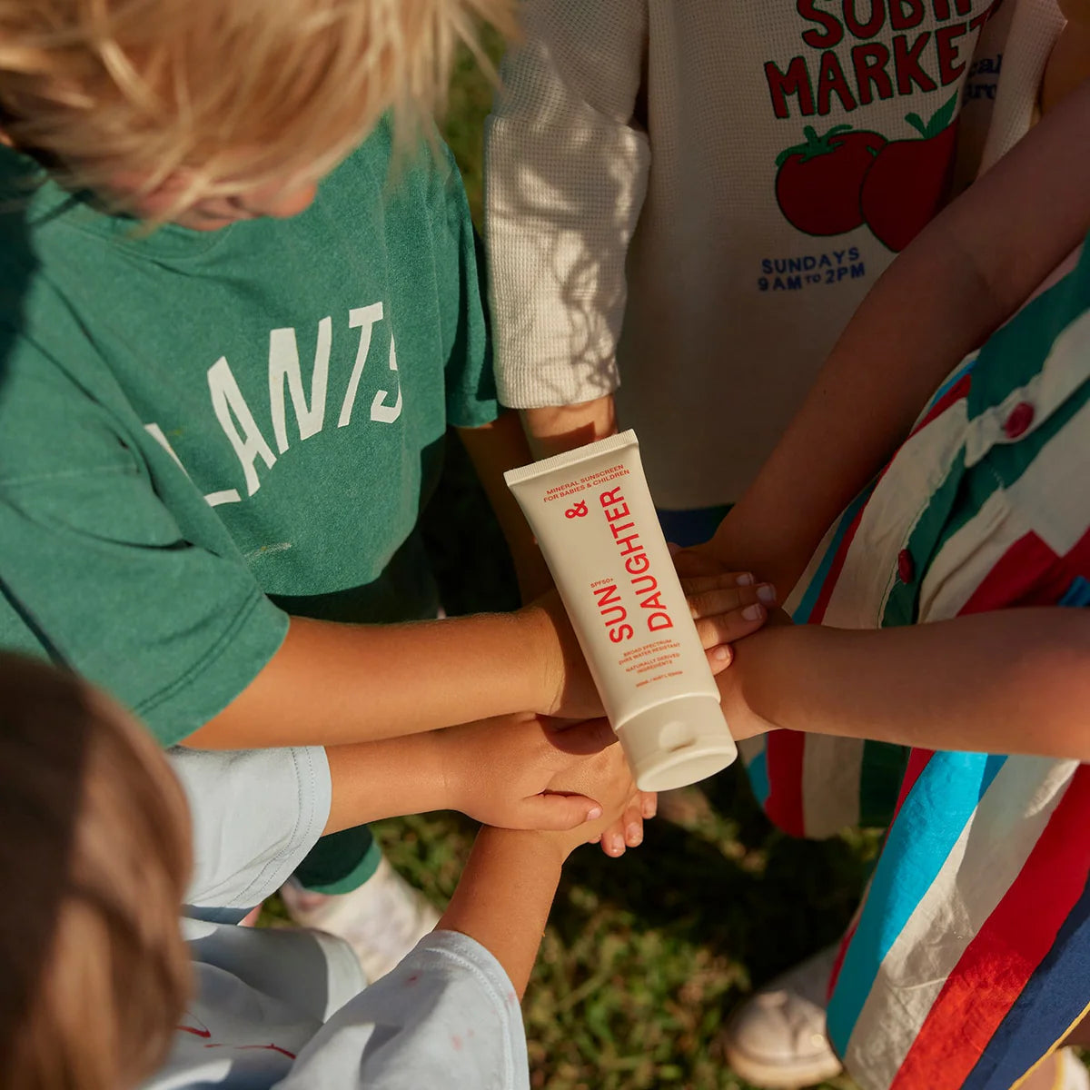 Children stand in a circle outdoors, each placing a hand on a Sun & Daughter SPF 50+ Mineral Sunscreen tube. They wear colorful clothing, including "Plants" and "Sunday Market" tops with tomato graphics, embracing the reef-friendly fun.