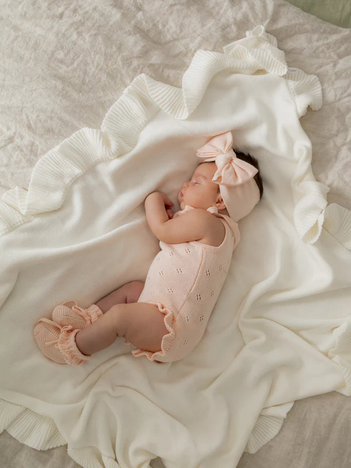 A sleeping baby wearing a light pink ZIGGY LOU Frill Bodysuit Primrose, matching headband with a large bow, and soft booties lies on a ruffled white blanket. The 100% cotton ensemble ensures the baby is comfortably resting on their side.