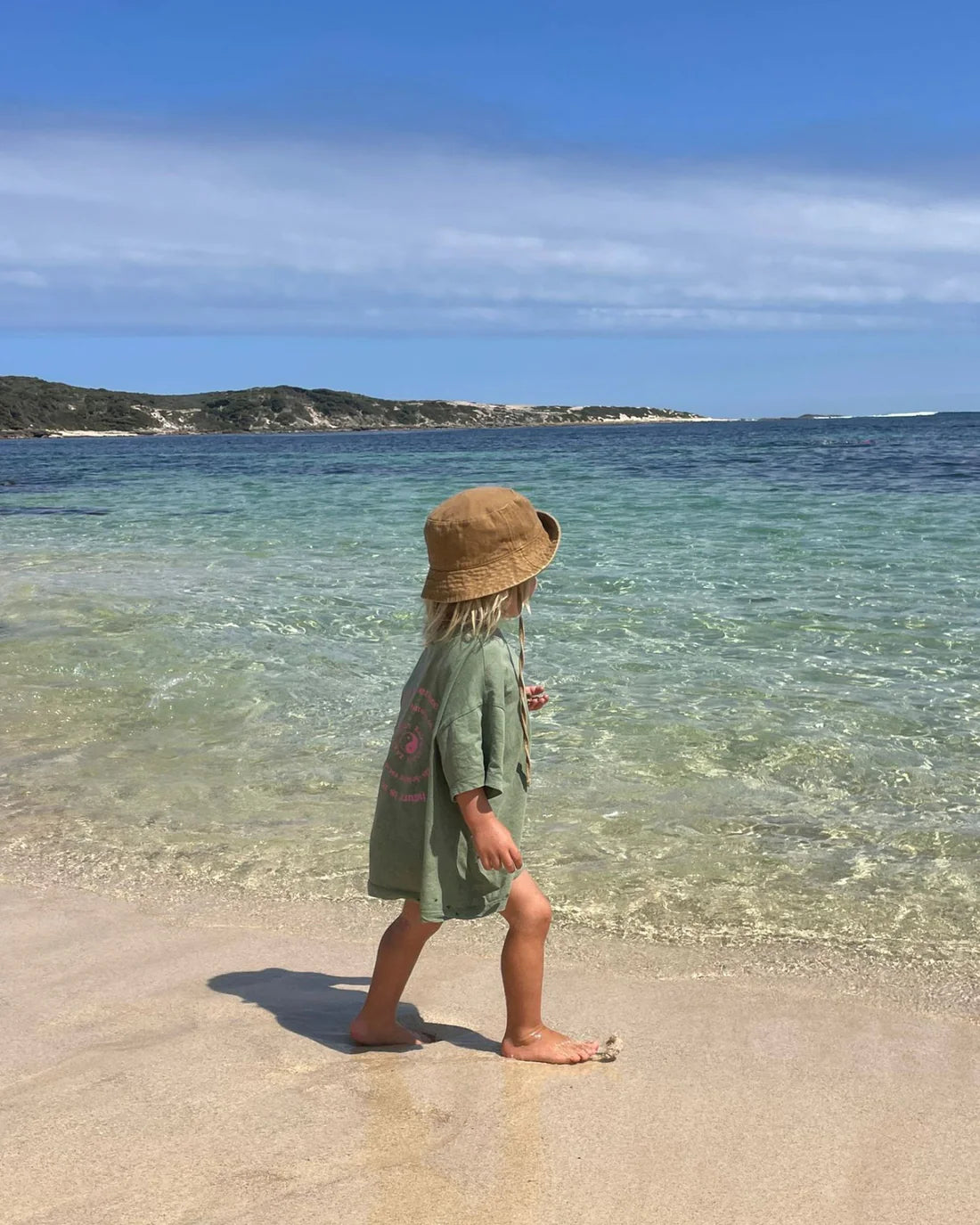 A child with long hair, wearing a ZIGGY ZAZA Ziggy Zaza ~ Love Your Mother Tee Lizard in an oversized fit and a brown sun hat, walks barefoot on the beach towards the clear water. The sky is blue with some clouds, and hills are in the background.