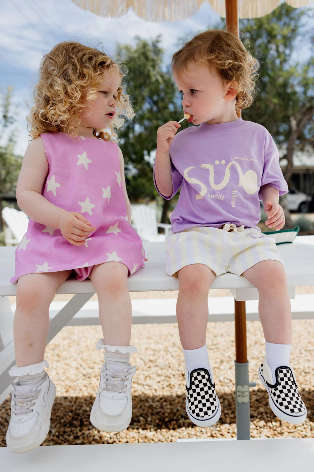 Two young children sit on a white bench outdoors. One wears a pink dress with white stars, and the other sports an Il Sole Tee in purple/cream by SUNDAY SIBLINGS, featuring an oversized fit and soft cotton fabric. Both have curly hair and casual shoes.