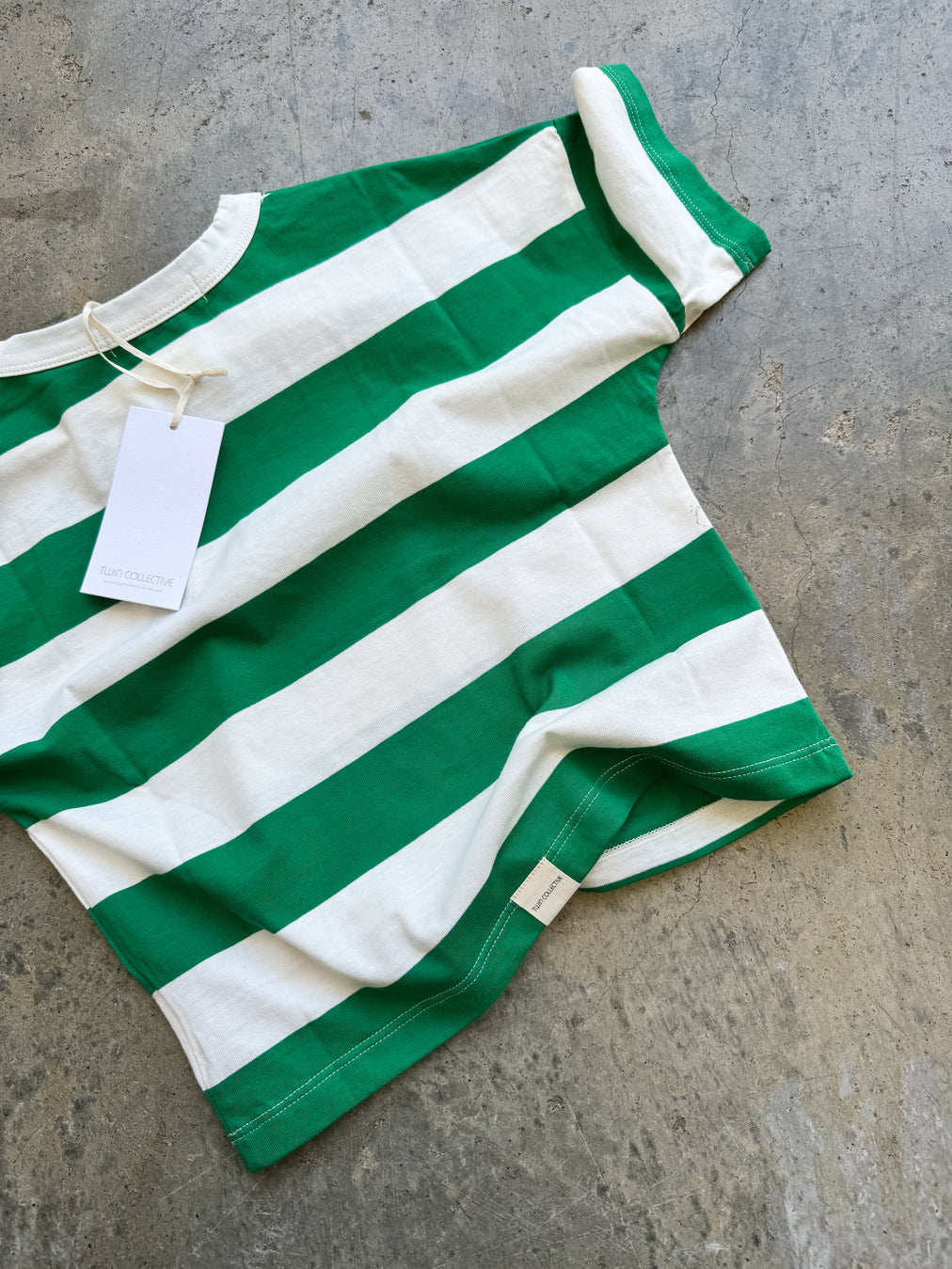 The Twin Collective ~ Stripe Tee Green, by TWIN COLLECTIVE, is a 100% cotton green and white striped T-shirt with a tag attached, spread out on a textured gray surface.