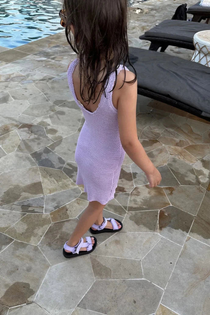 A child with wet hair stands by a pool wearing a light purple dress and stylish white sandals. The Olympia Velcro Sandal Lilac ensures comfort as they walk on stone tiles near black lounge chairs, proudly showcasing the TINY TROVE logo and its flexible sole for playful steps.