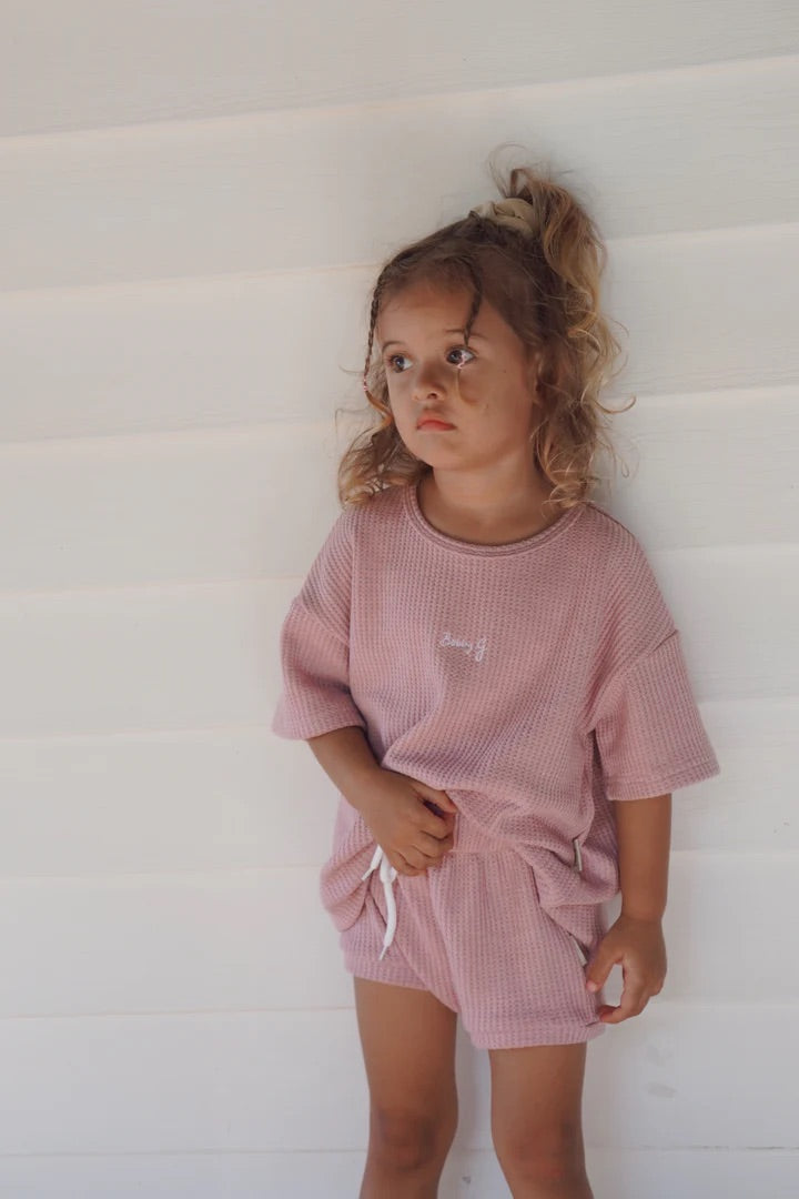 A young child with curly hair stands against a white wall, wearing the Coby Shorties Dusty Rose by BOBBY G BABY WEAR. The matching outfit, made from soft waffle fabric with a stretchy waistband, complements the child's neutral expression as they gaze to the side.