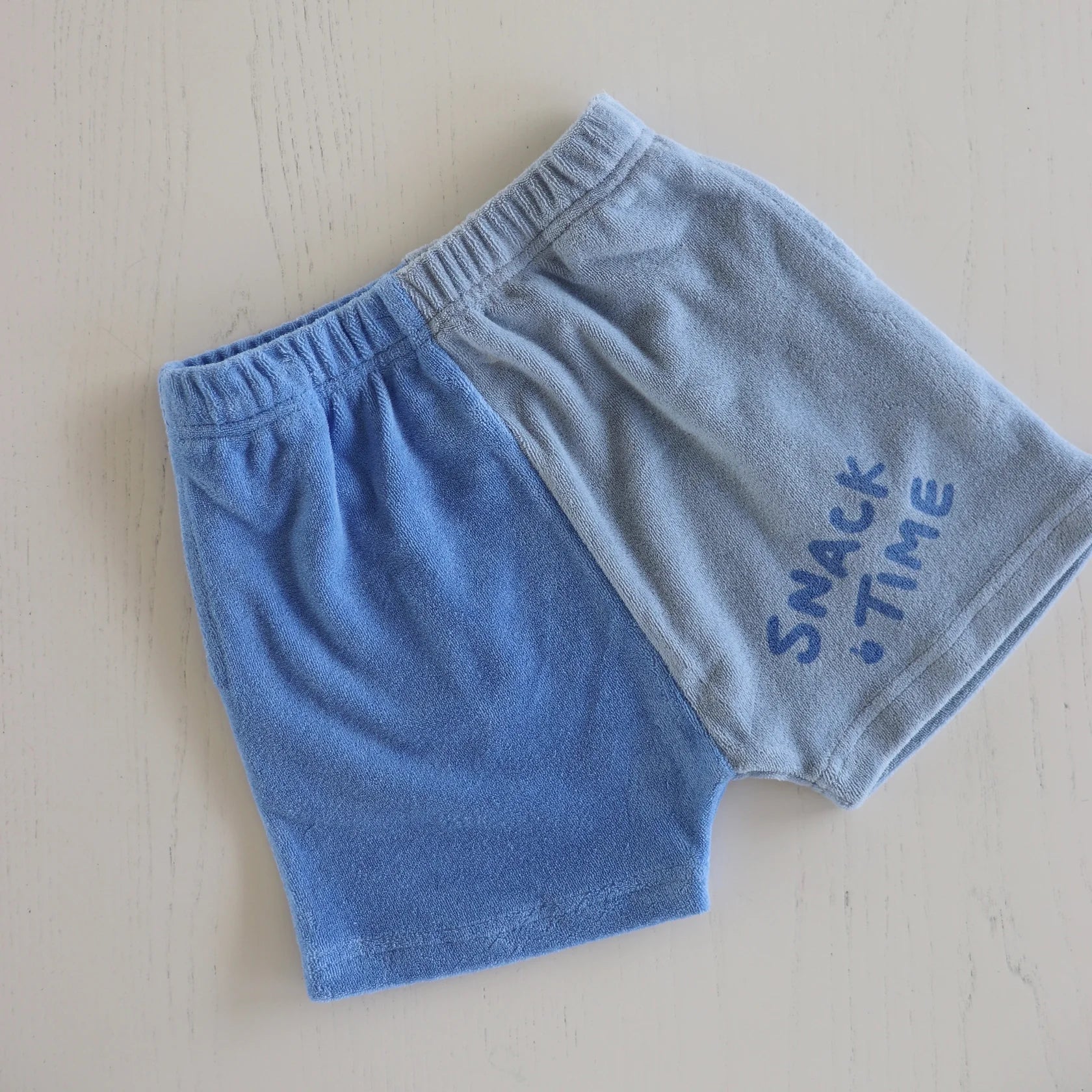 A pair of GOLDEN CHILDREN's Play Lounge Shorts Pool for children lays flat on a white surface. Made from terry towel cotton, they feature an elastic waistband and are color-blocked with one side light blue and the other darker blue. The light blue side is adorned with a "SNACK TIME" print in dark blue.