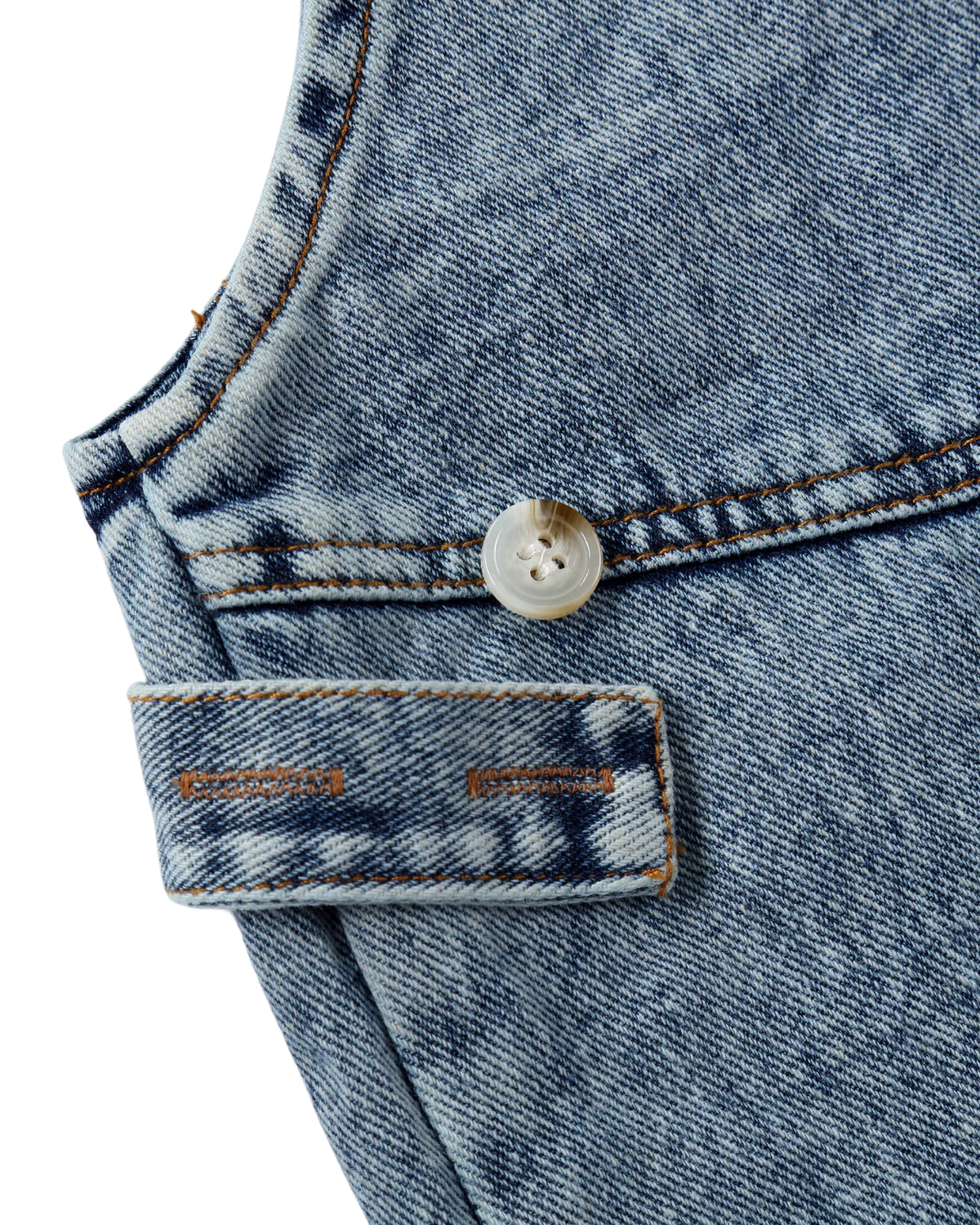 Close-up of the SUSUKOSHI Denim Overalls Sky Blue featuring a white button, an orange rectangular label with unreadable text, and contrasting stitching along the pocket and seams. Made from organic cotton, the fabric has a slightly faded, worn texture.