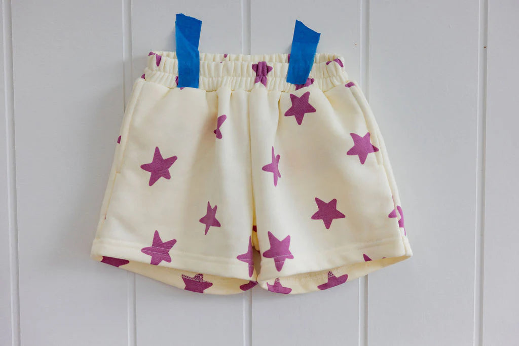 A pair of Sunday Siblings Kiddo Shorts in cream with pink stars is taped to a white paneled wall with blue tape. Made from soft French terry cotton, these children's shorts feature an elastic waistband.