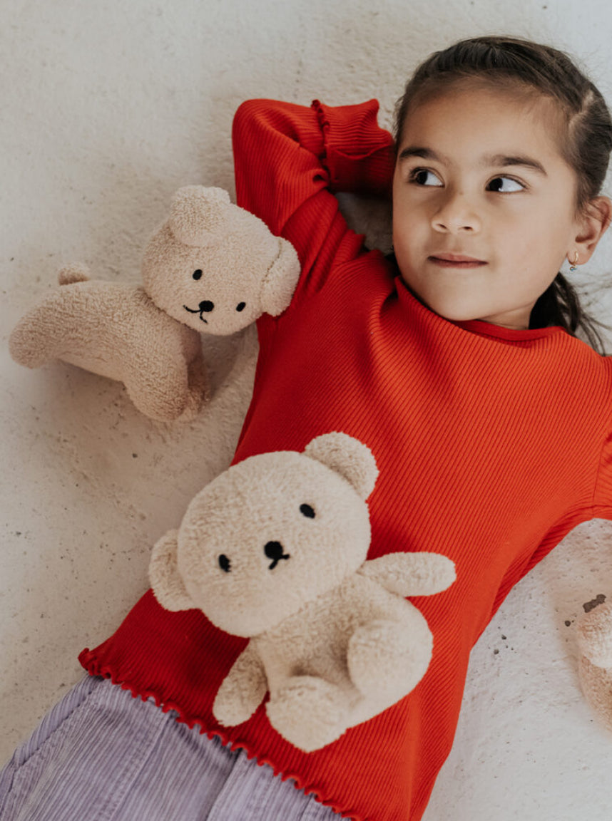 A young girl with dark hair lies on a light-colored, textured surface. She is wearing a bright red sweater from the Miffy Terry Collection and cuddling two fuzzy, light brown teddy bears. One of the bears is the "Boris Bear Sitting Terry Beige 23 cm" from BON TON TOYS X MIFFY. She looks up with a calm expression, her body relaxed.