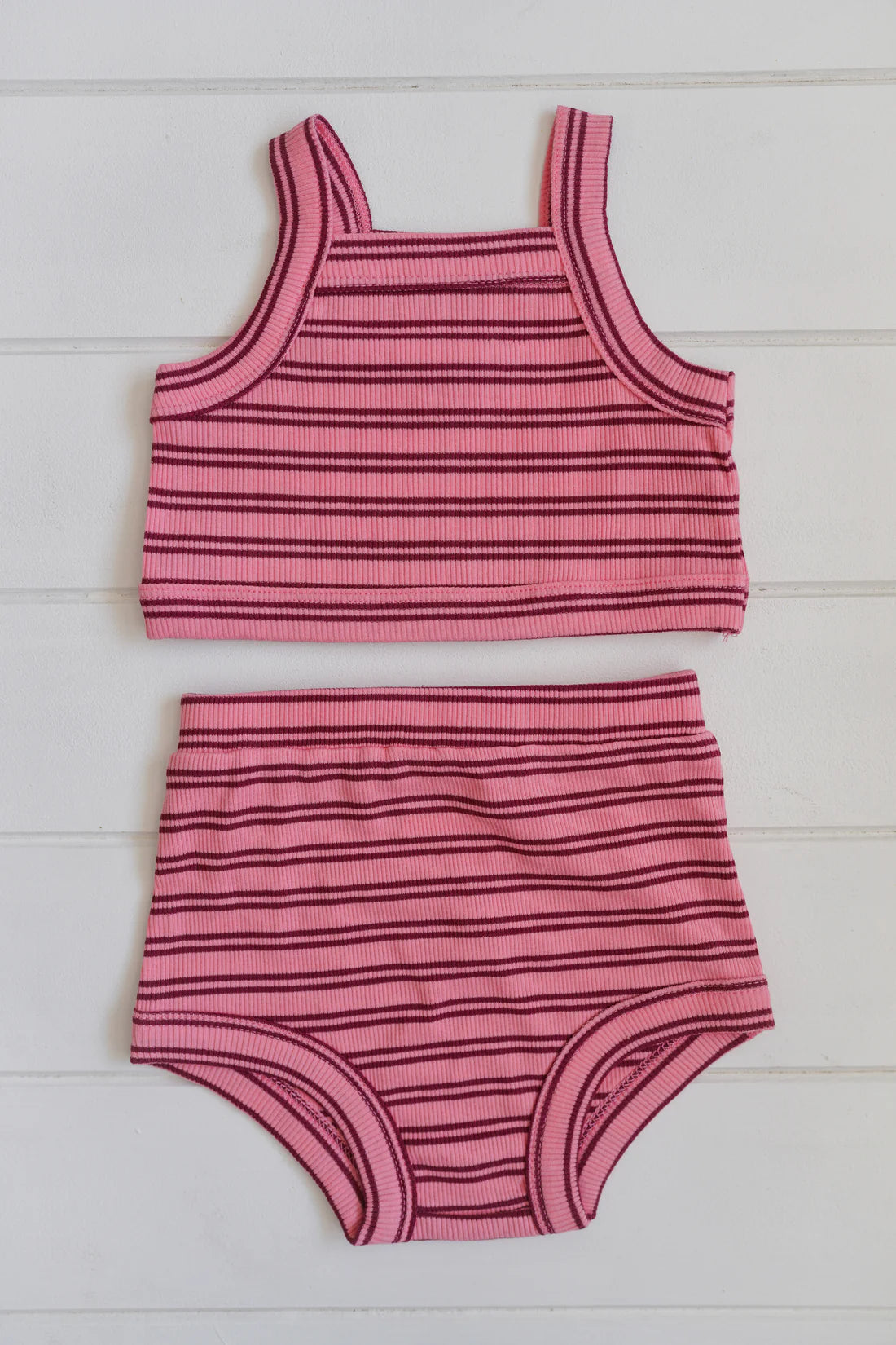 An engaging set for toddlers, the Cleo Rib Set by MILKY DESIGNS showcases a pink swimsuit with red horizontal stripes, crafted from ribbed cotton. This set includes a sleeveless top and high-waisted bottoms, both stylishly displayed flat on a white wooden surface.