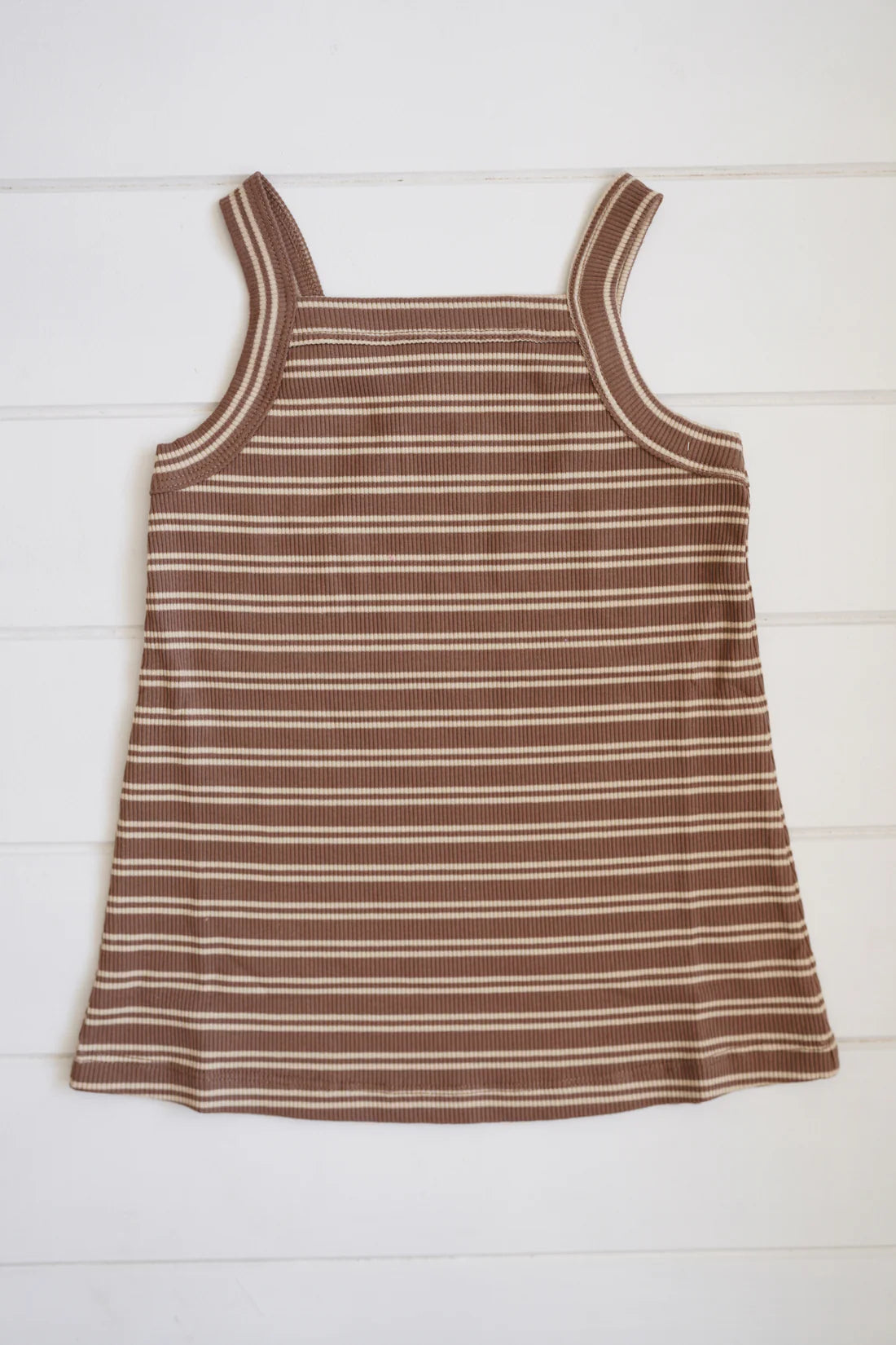 The Bobbi Rib Dress Choc Stripe by MILKY DESIGNS, a sleeveless ribbed cotton dress featuring brown and white stripes and a square neckline, is laid flat against a light wooden background.