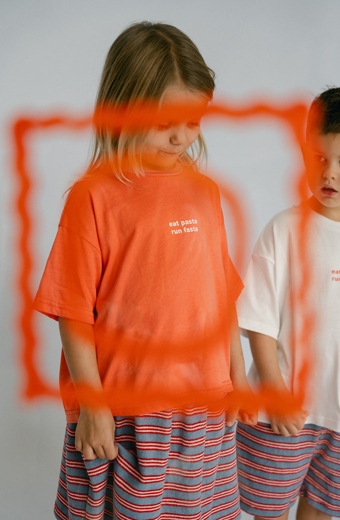 A young kid wearing the Lenn pasta tee and a striped skirt standing next to a boy.