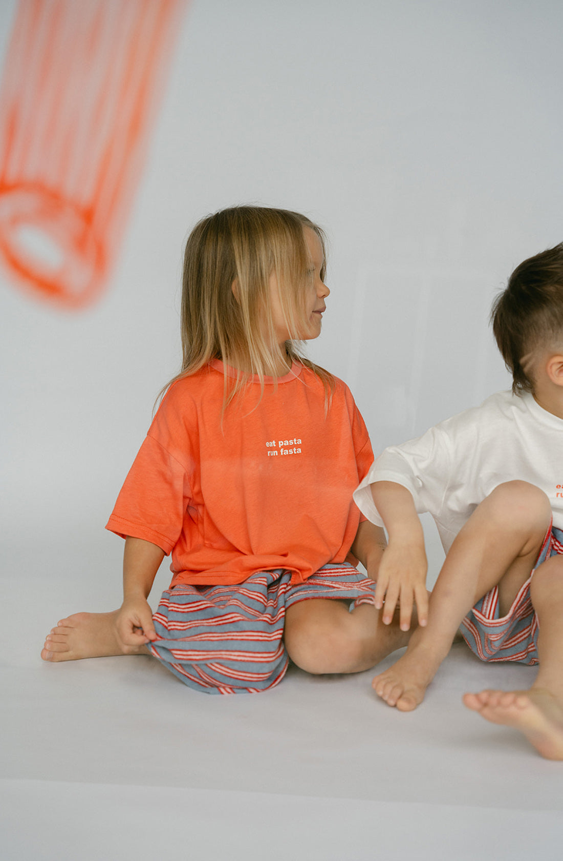 A young kid wearing the Lenn pasta tee with a striped skirt sitting next to a young boy