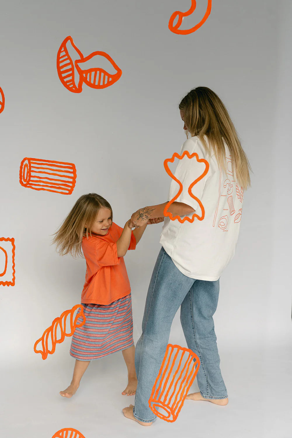 A young child in an orange shirt and striped skirt playfully dances with an adult wearing the Women's Pasta Tee White by LENN LABEL paired with jeans. Orange pasta-shaped drawings on a light background surround them, adding whimsy to their joyous moment.