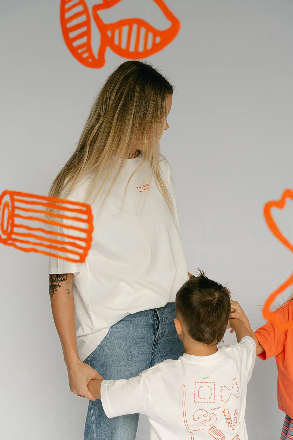 A woman wearing the LENN LABEL Women's Pasta Tee in white, styled in a lightweight and oversized fit, pairs it with jeans while holding the hand of a child dressed in a matching cotton jersey. Another individual is partially seen nearby against a backdrop decorated with vibrant orange illustrations of various objects.