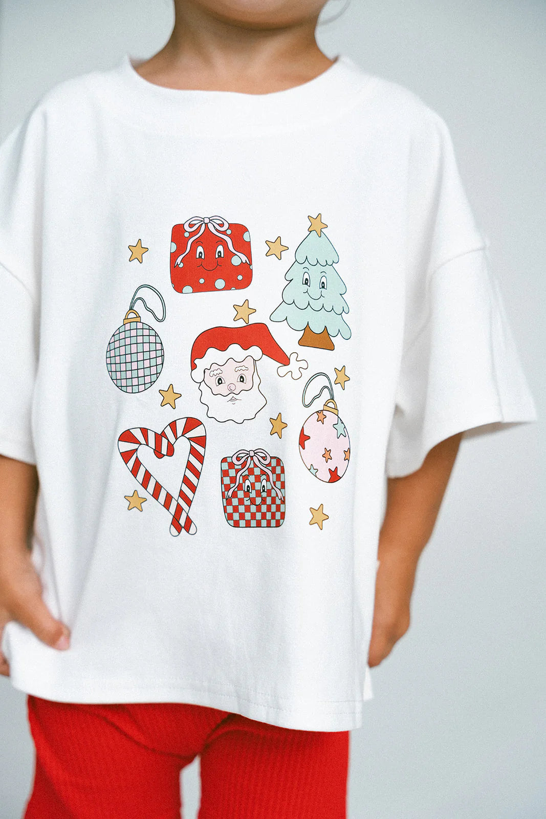 A child is gleefully wearing the Merry & Bright Icons Oversized Tee by MYLA JANE, featuring charming Christmas-themed illustrations such as Santa, a tree, presents, candy canes, ornaments, and stars. Ideal for holiday season festivities and lively Christmas parties, this festive outfit is complemented by cheerful red pants.