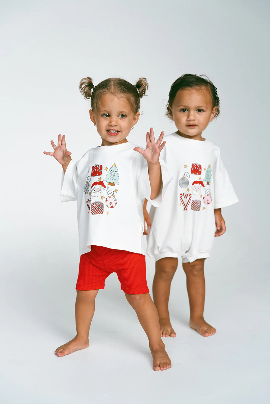 Two toddlers stand side by side wearing the Merry & Bright Icons Oversized Tees from MYLA JANE, featuring delightful Christmas-themed designs. The child on the left sports red shorts with pigtails, and the child on the right complements their look perfectly. Both are barefoot and beaming, encapsulating the joy of the holiday season against a simple backdrop.