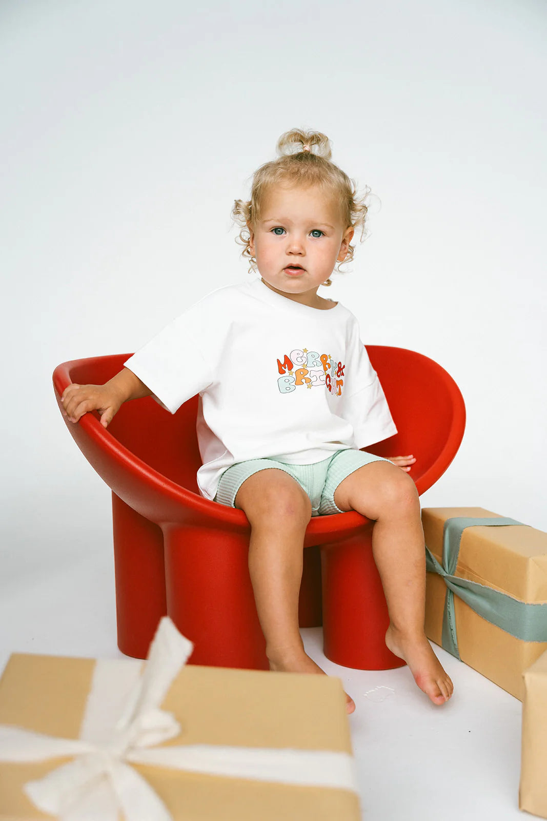 A young child with curly hair is sitting on a modern, red chair, wearing an oversized tee from the Merry & Bright Logo collection by MYLA JANE and green shorts. Two wrapped gift boxes with ribbon are positioned in front of the chair, adding a touch of the holiday season spirit. The background is plain white.