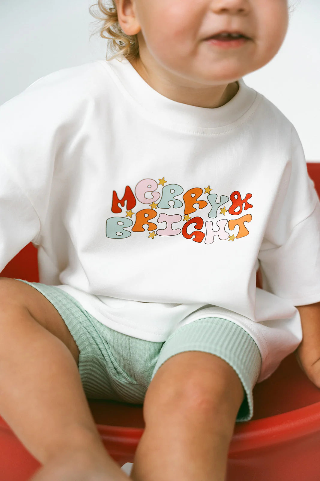 A young child sits wearing the MYLA JANE Merry & Bright Logo Oversized Tee, capturing the holiday season spirit with its festive design. They are on a red surface, their light hair partially visible, and sporting pastel-colored shorts that complement the joyous theme.