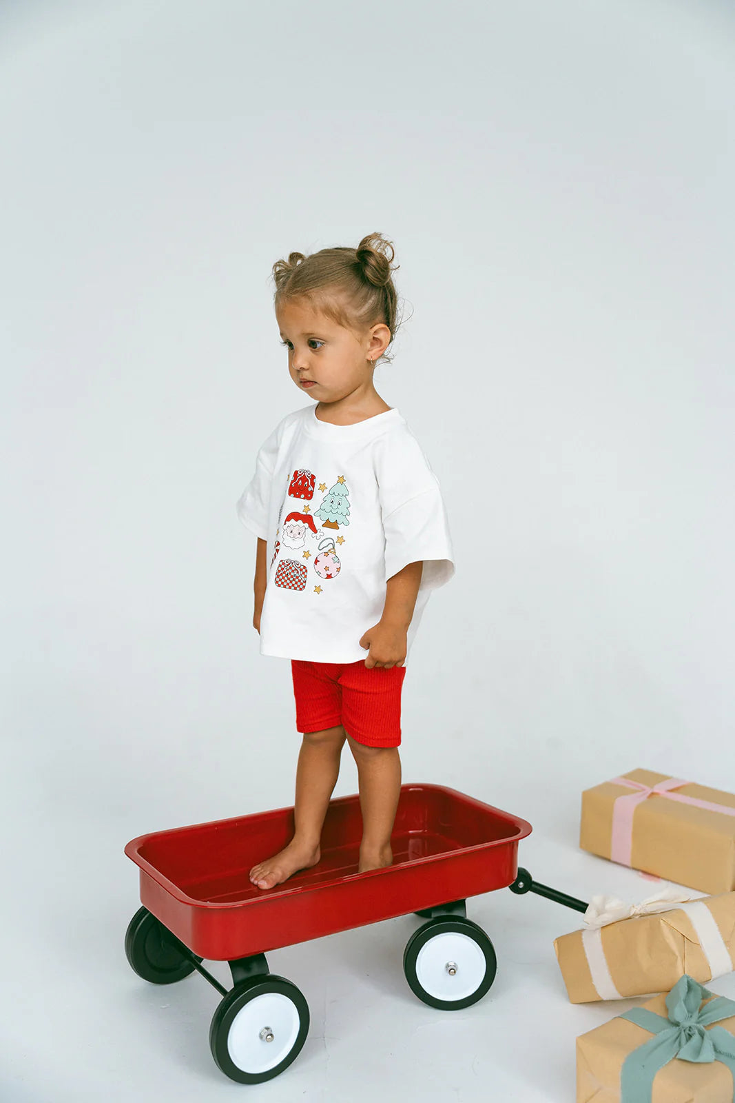 A young child with two small buns stands inside a red wagon wearing the Merry & Bright Icons Oversized Tee by MYLA JANE, paired with red shorts. The plain background and wrapped gifts evoke the holiday season, capturing a Merry & Bright vibe nearby.