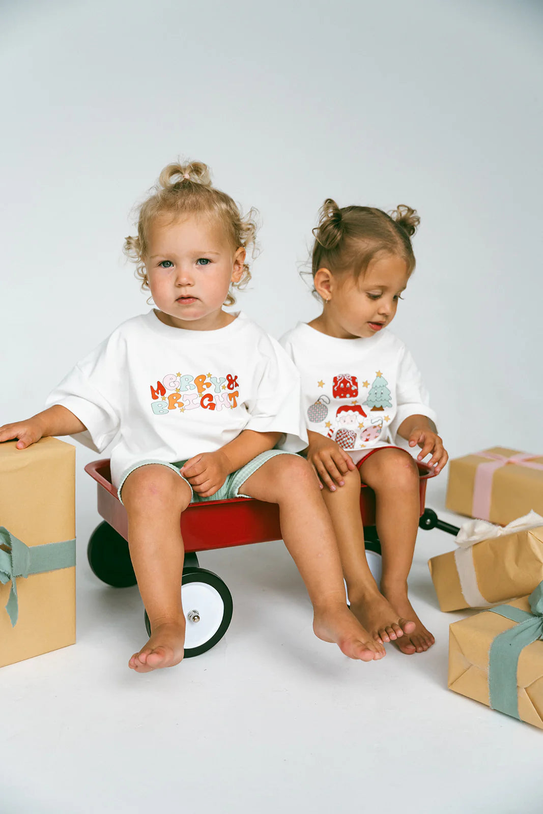 Two young children sit in a red wagon surrounded by gift boxes, capturing the joyful spirit of the holiday season. They wear festive T-shirts—one features the Merry & Bright Logo Oversized Tee from MYLA JANE while the other is adorned with holiday-themed illustrations. With light hair and bare feet, they create a cozy atmosphere.