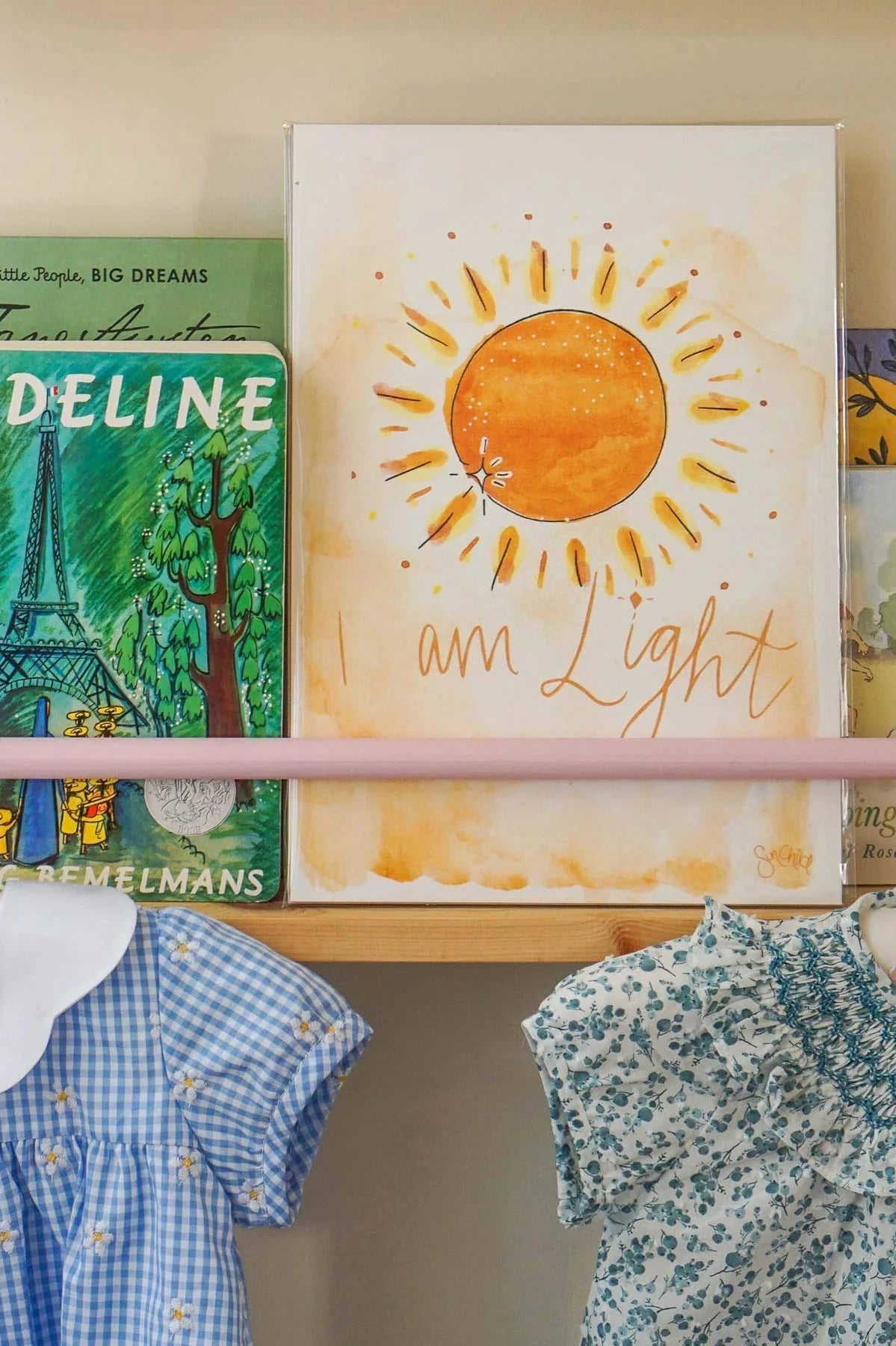 A shelf displays an A4 unframed SunChild "I Am Light" Art Print, showcasing a sun and the affirmation "I am Light." Alongside it are kids' affirmation wall art and children's books, including one with an Eiffel Tower cover. Below, a rod holds two children's dresses featuring floral and checkered patterns.